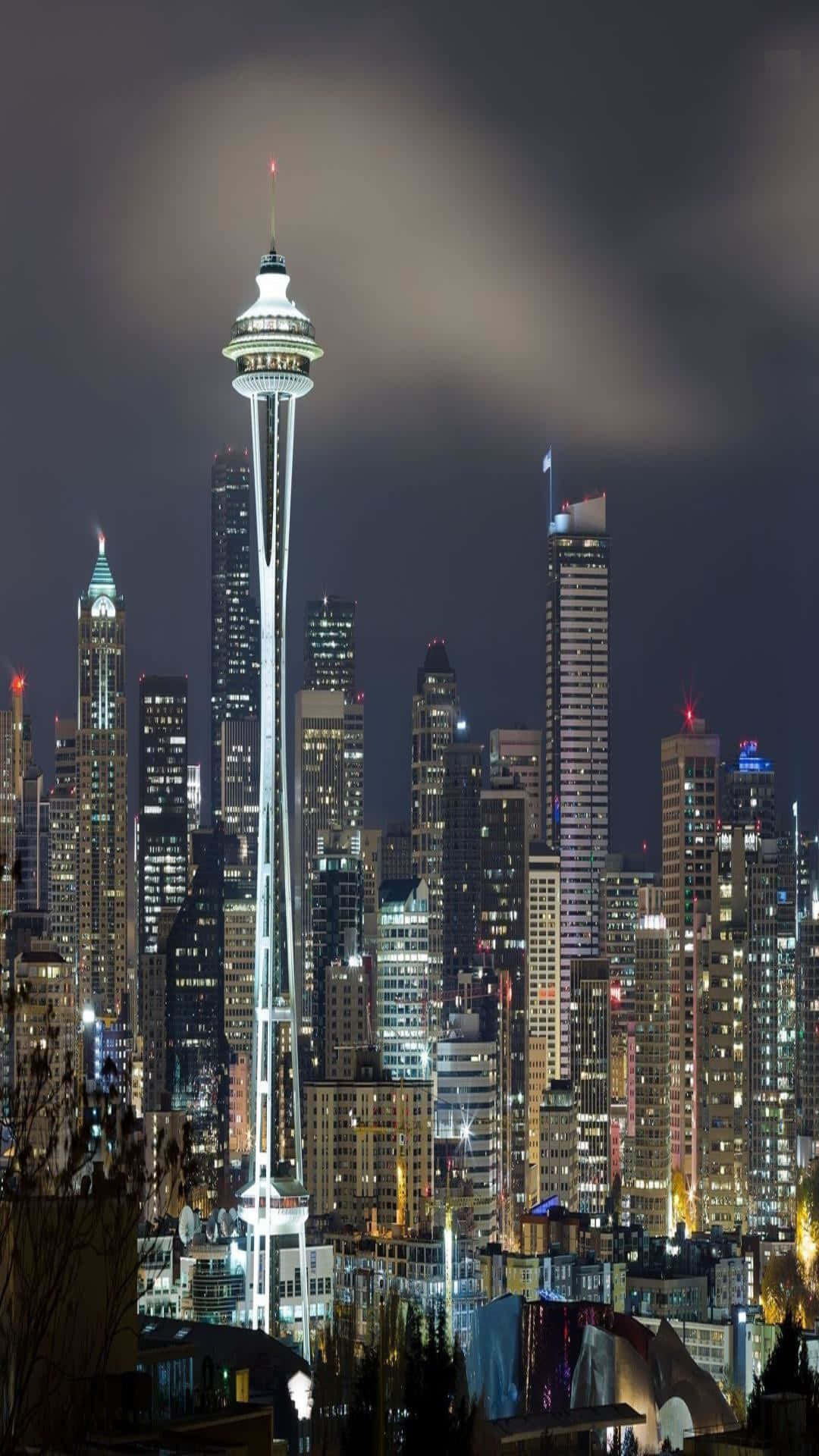 washington state 桌布 壁紙 — 西雅圖的太空針塔在黑暗多雲的夜空下，在密集的摩天大樓城市景觀上方明亮發光。