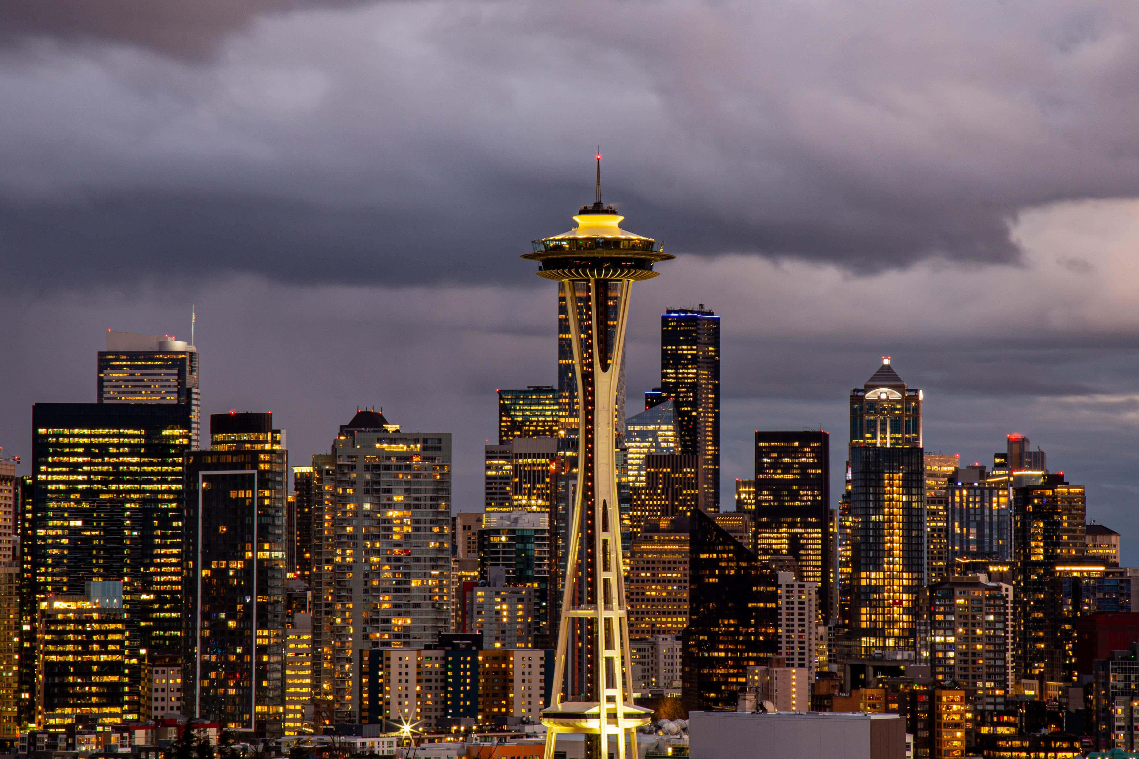Seattle At Night 桌布 壁紙 — 西雅圖黃昏天際線，太空針塔在烏雲中閃耀，摩天大樓的燈光閃爍。