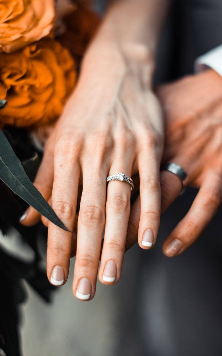 Wedding Rings 桌布 壁紙 — 親密特寫兩隻手緊握在一起，帶有閃爍的鑽石訂婚戒指和結婚戒指，背景是溫暖的橙色玫瑰。