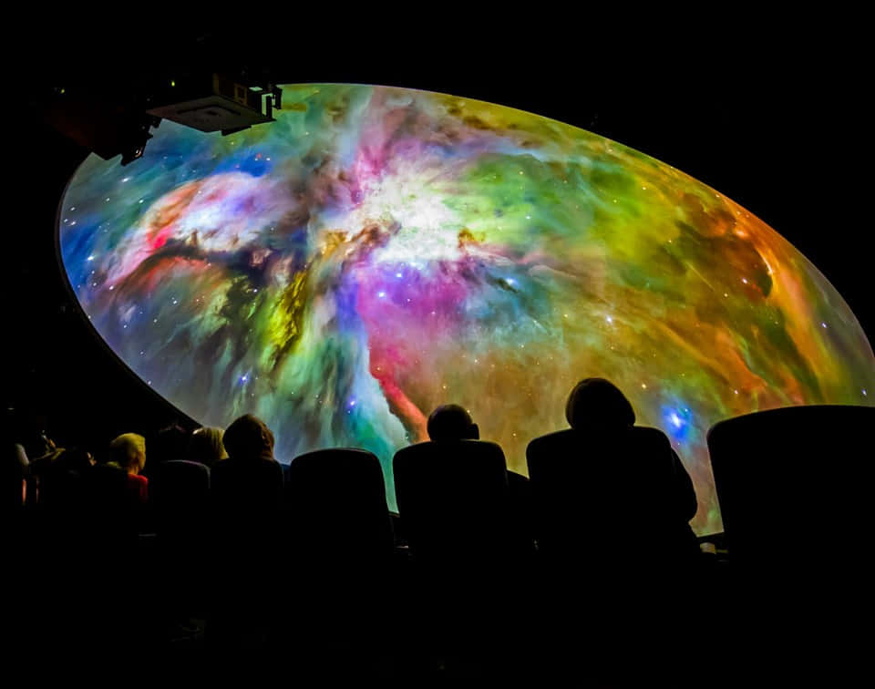 Planetarium 桌布 壁紙 — 觀眾剪影仰望巨大穹頂投影，呈現出彩虹色調的鮮豔宇宙星雲。