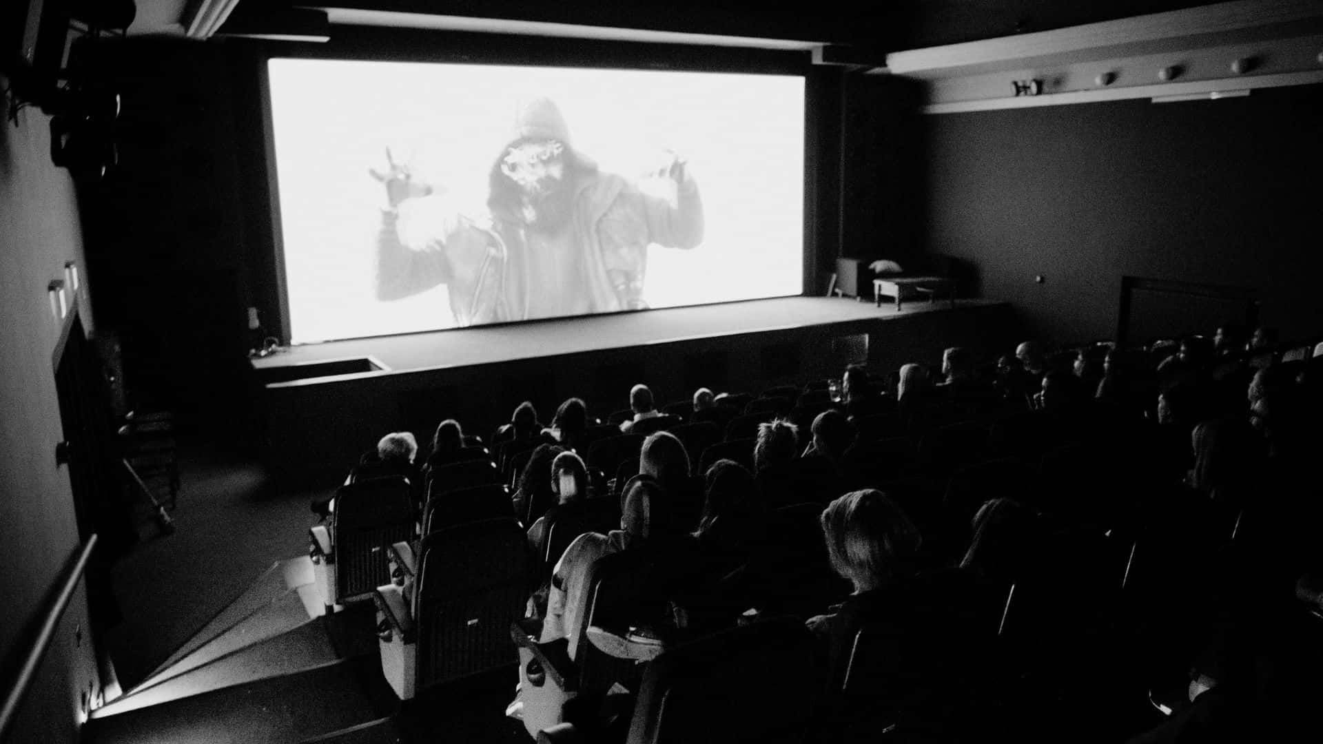 Black And White Movie 桌布 壁紙 — 觀眾在昏暗的影院中觀看螢幕上一個戴著兜帽的人物，畫面呈現強烈的黑白對比。