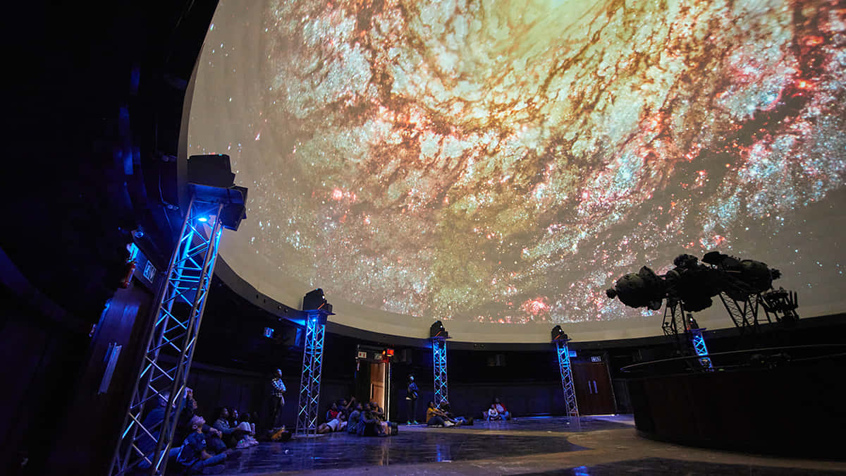 Planetarium 桌布 壁紙 — 觀眾坐在一個巨大的圓頂下，圓頂展示著充滿活力的宇宙星雲，有旋轉的紅色、金色和藍色。