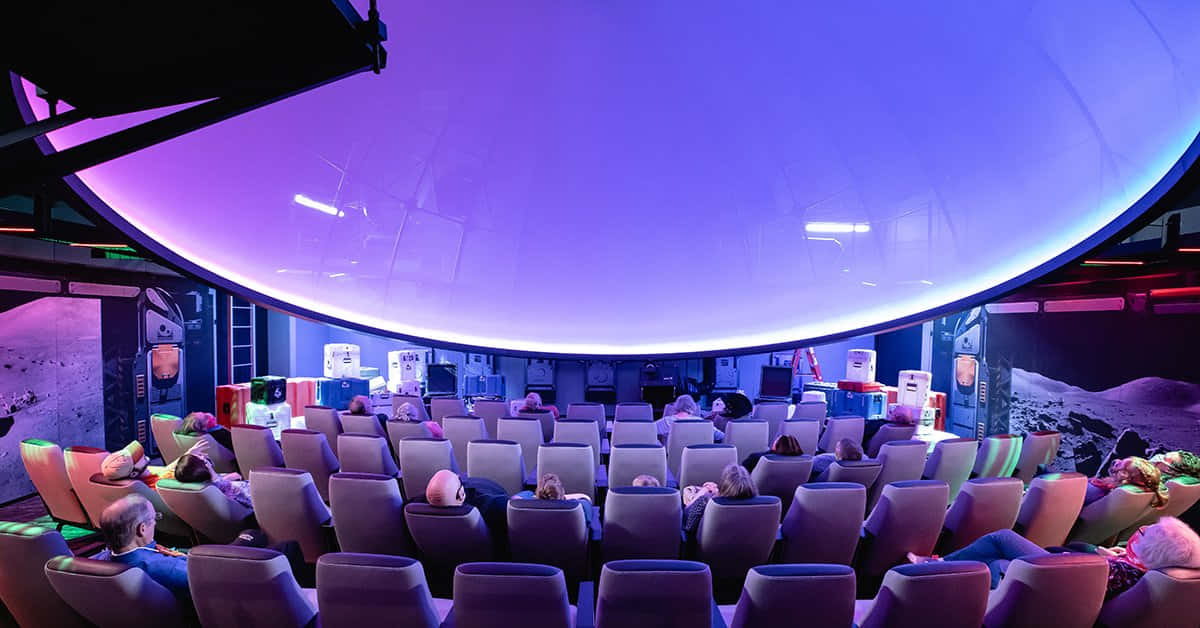 Planetarium 桌布 壁紙 — 觀眾坐在未來風格的圓頂劇院中，頭頂是發光的紫色天花板，搭配沉浸式天文館視覺效果。