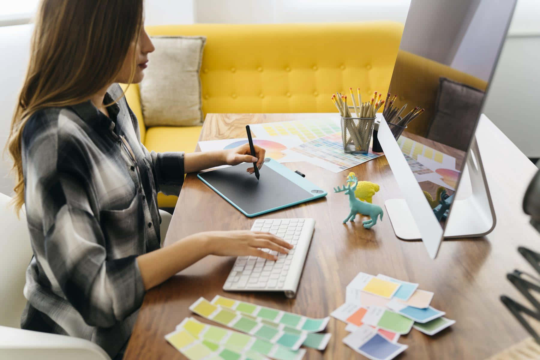 graphic design 桌布 壁紙 — 設計師在木桌上使用繪圖板、色板，背景有黃色簇絨沙發。