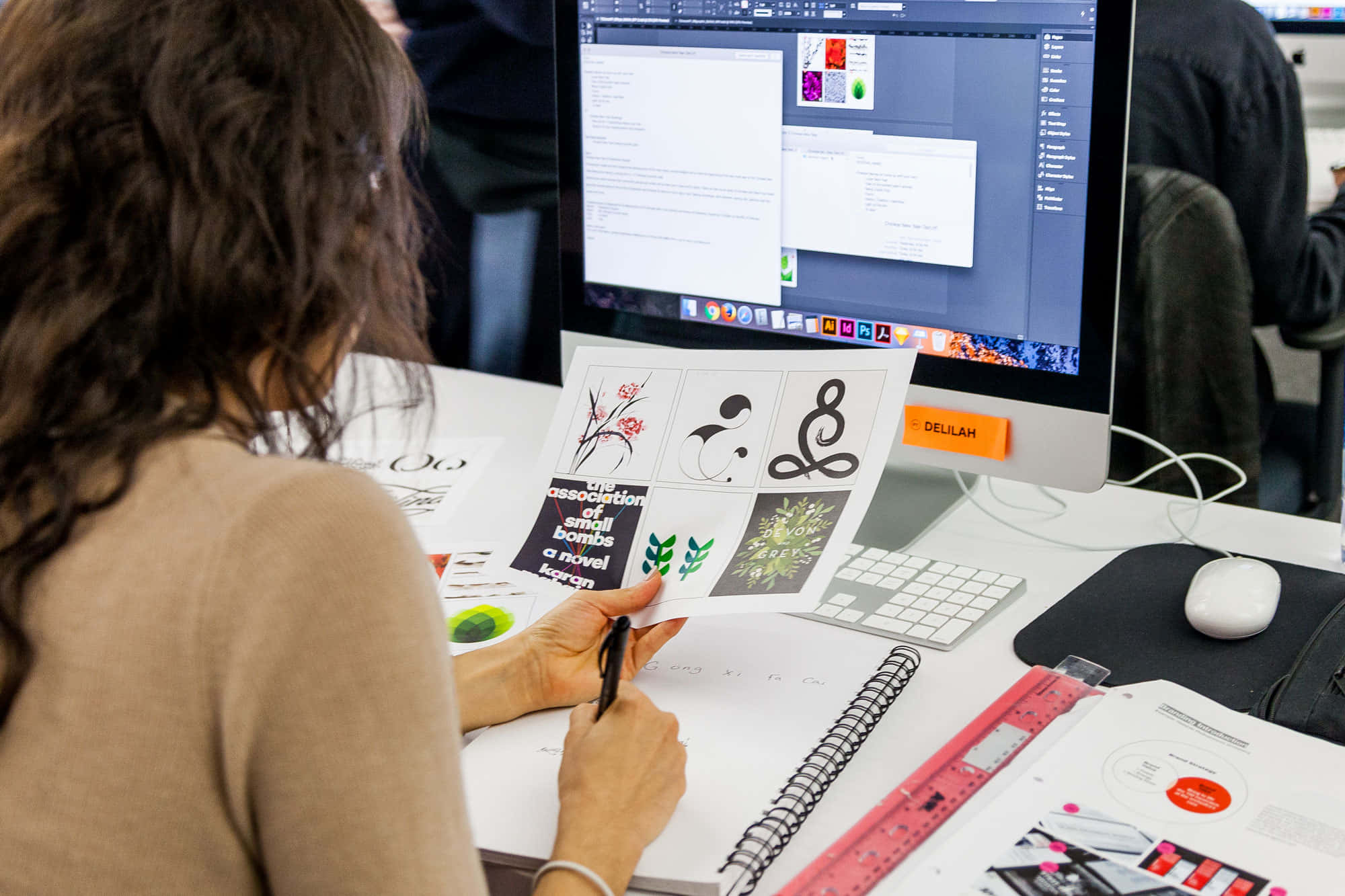 graphic design 桌布 壁紙 — 設計師正在一張雜亂的桌子上工作，桌上有一台顯示設計軟體的電腦顯示器，同時審閱紙上的圖形概念。