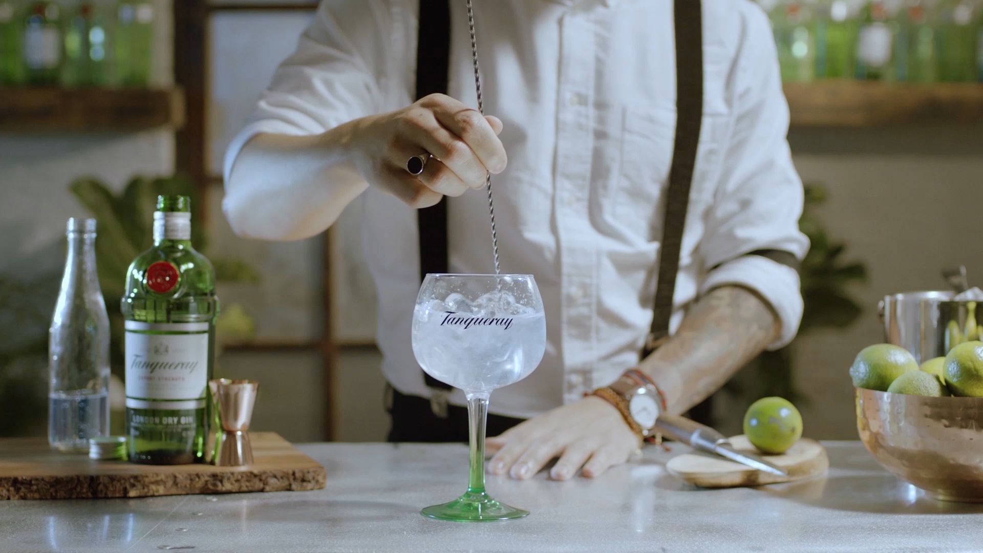 bartender 桌布 壁紙 — 調酒師在一個鄉村風格的酒吧檯上，用一個品牌玻璃杯攪拌琴酒雞尾酒，周圍有萊姆和酒瓶。