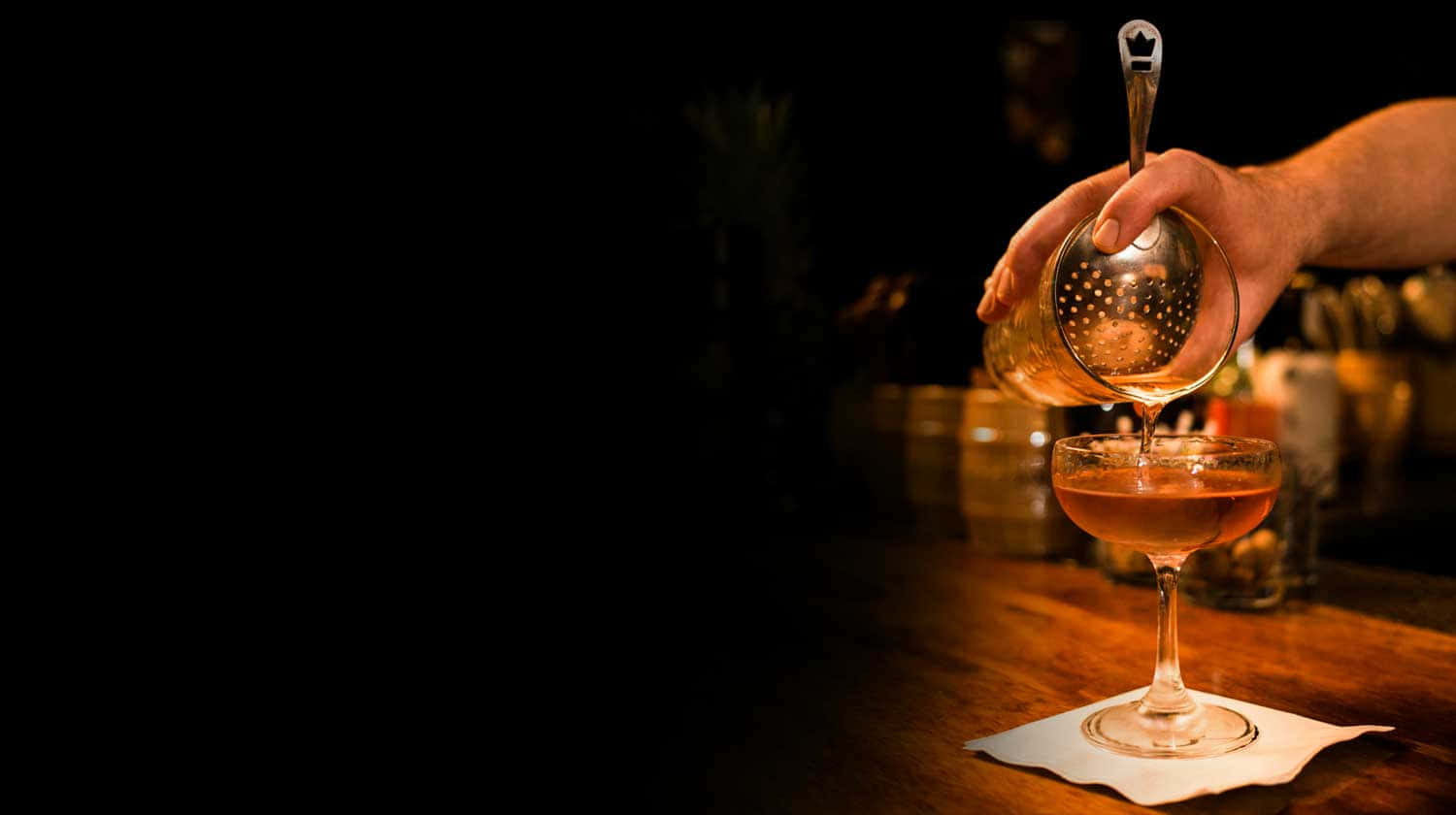 alcohol 桌布 壁紙 — 調酒師在夜晚透過隔渣器將金色雞尾酒倒入木吧台上的庫佩酒杯中。