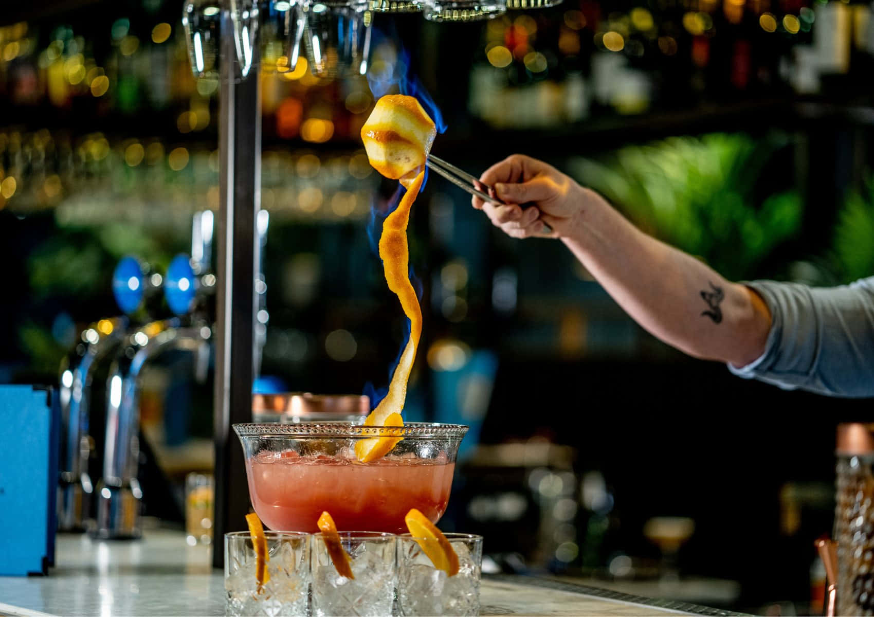 alcohol 桌布 壁紙 — 調酒師在昏暗的酒吧中，在充滿散景燈光的環境下，將橙皮點火燃燒在鮮豔的粉紅色雞尾酒上。