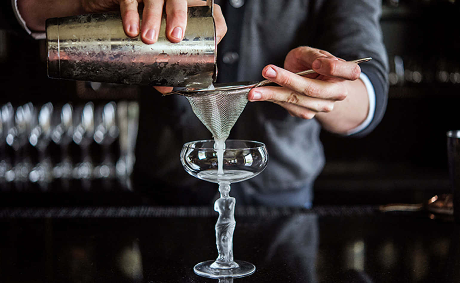 alcohol 桌布 壁紙 — 調酒師在深色吧檯上，用細濾網將清澈的雞尾酒倒入優雅的笛形杯。