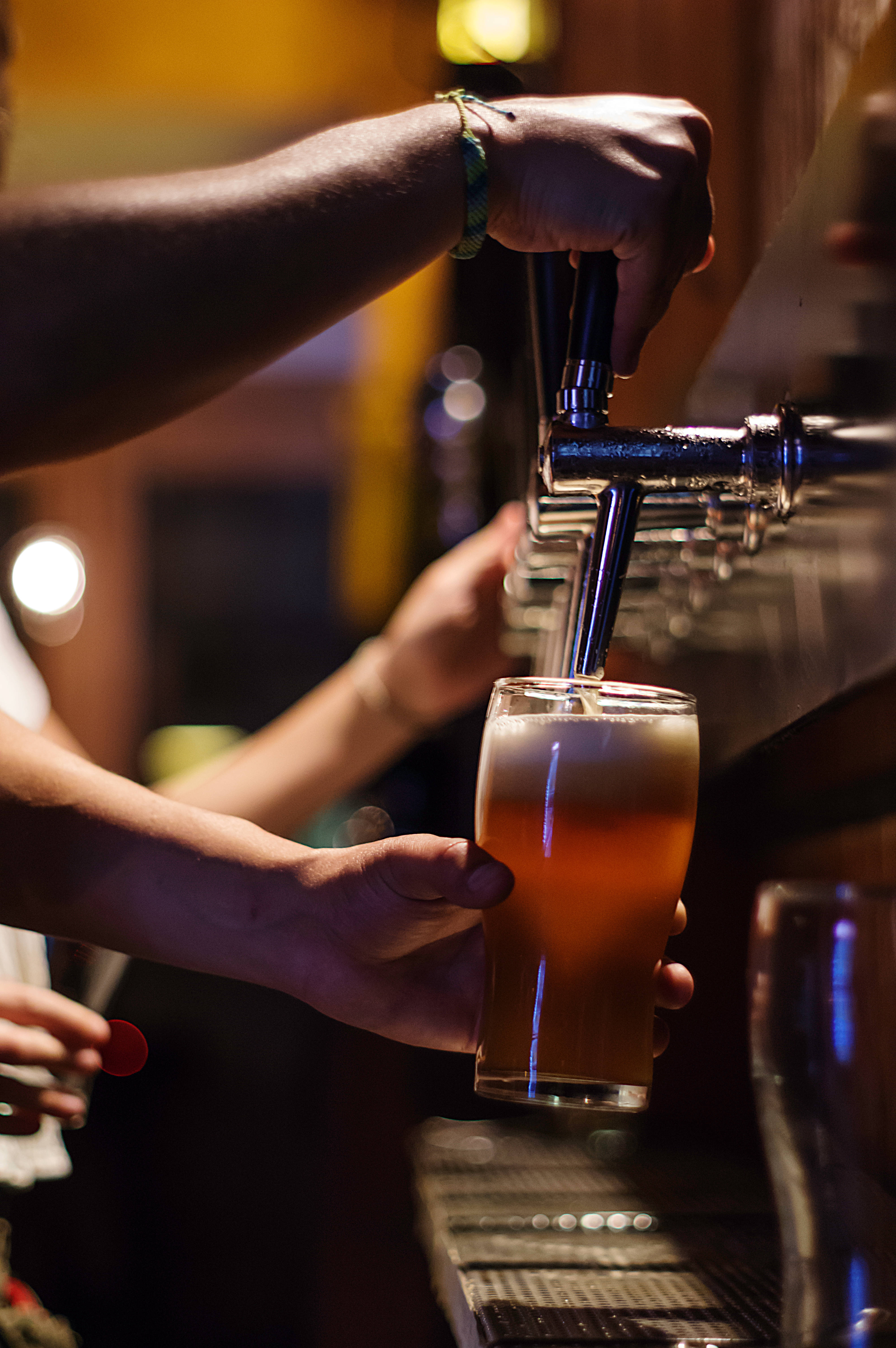 Beer 桌布 壁紙 — 調酒師在燈光昏暗、帶有溫暖金色照明的酒吧中，從水龍頭將帶有泡沫的琥珀色啤酒倒入玻璃杯。