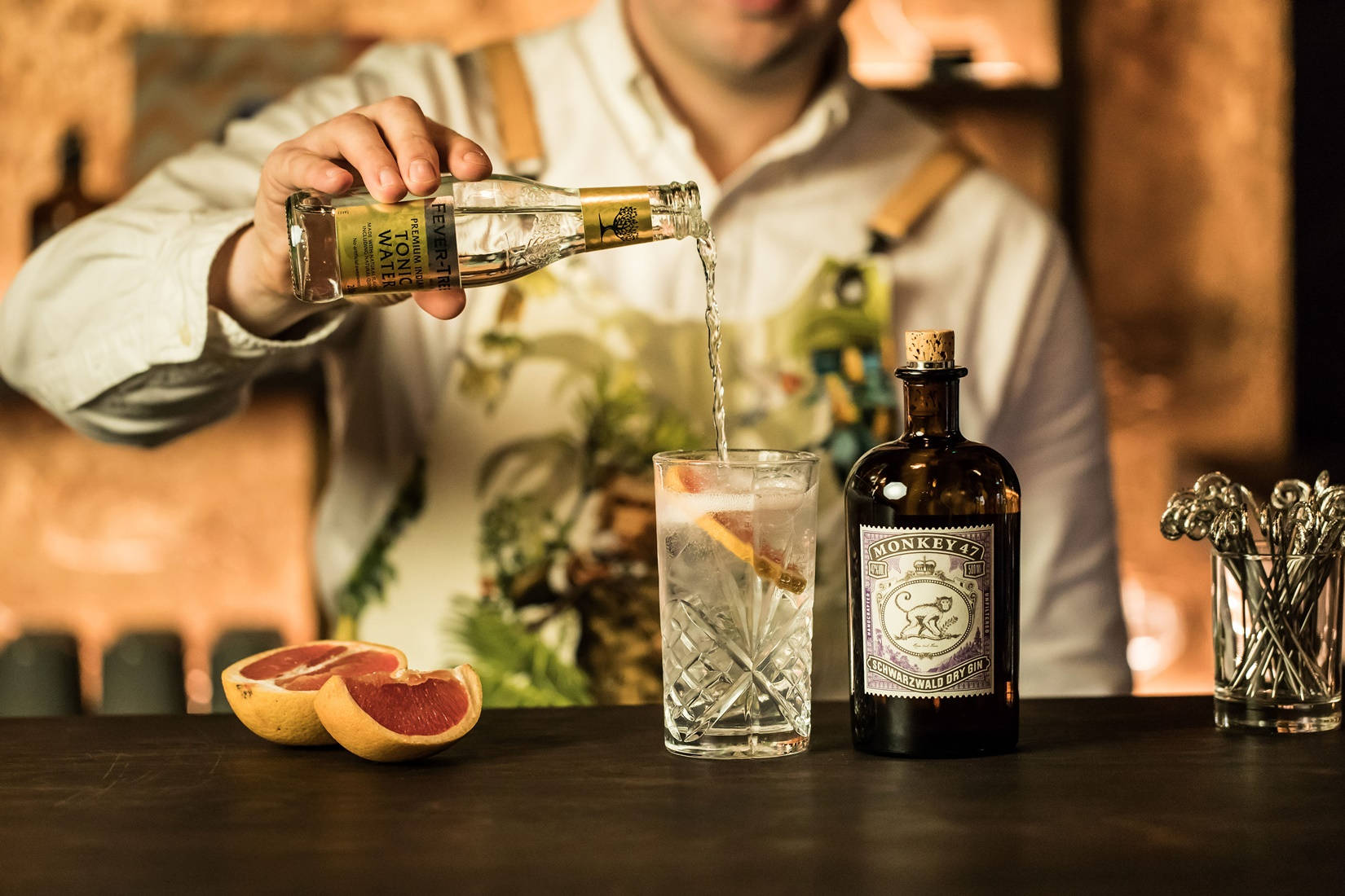 cocktail 桌布 壁紙 — 調酒師在黑暗的吧檯上，將通寧水倒入裝飾有柑橘和冰塊的琴酒雞尾酒杯中。