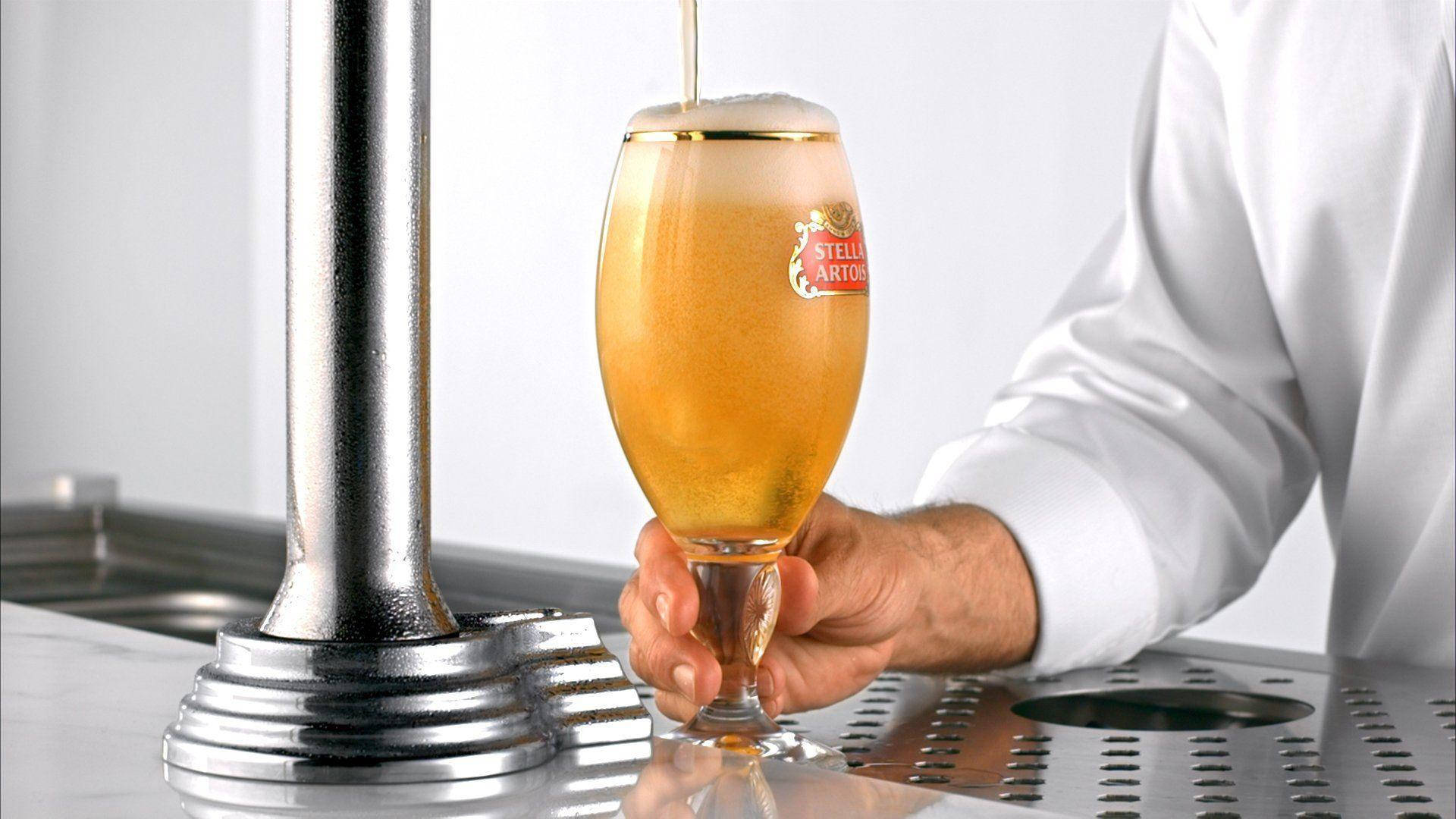 Drink 桌布 壁紙 — 調酒師將金黃色的 Stella Artois 啤酒倒入品牌玻璃杯中，帶有綿密泡沫，背景是乾淨的金屬吧檯。