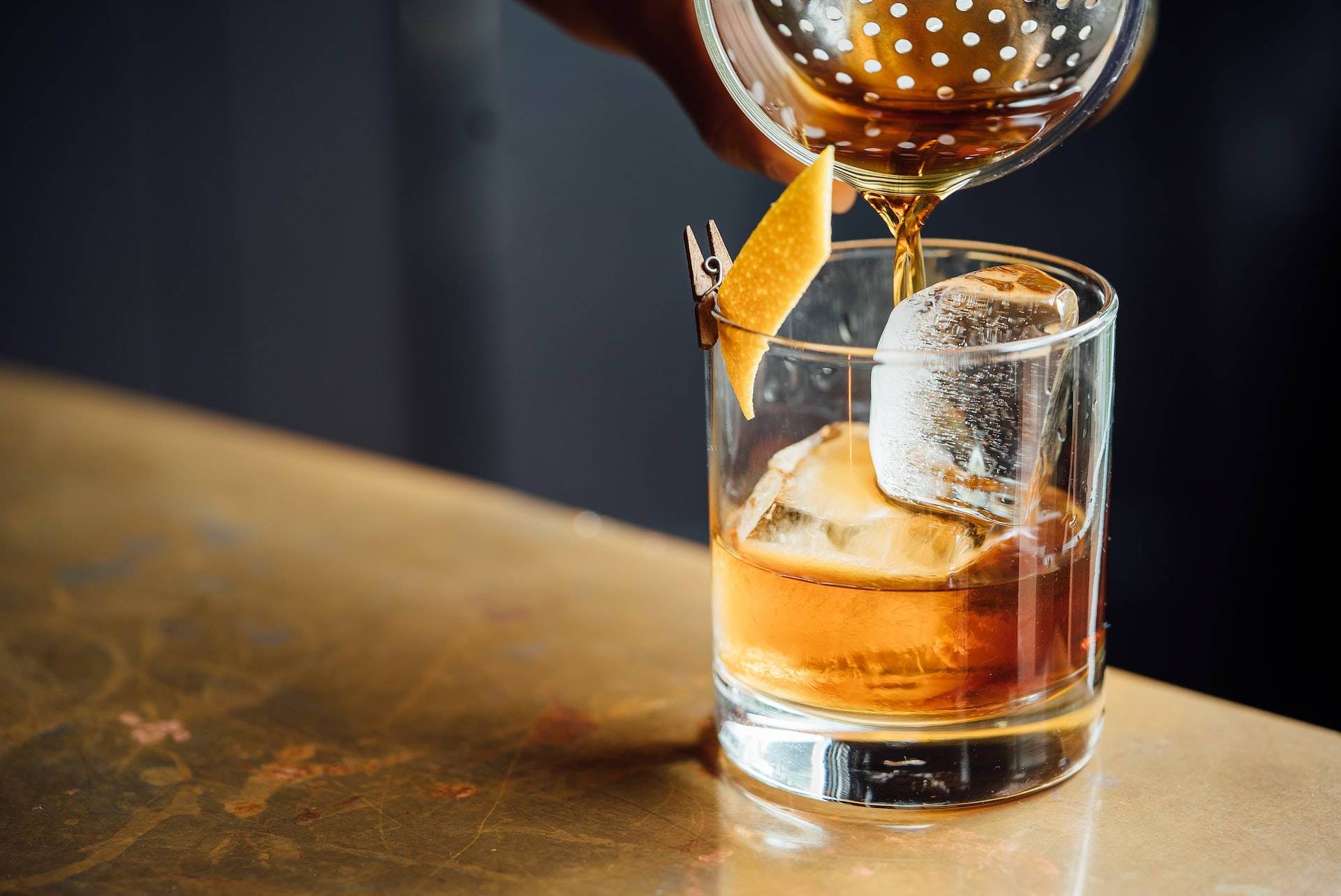 Drink 桌布 壁紙 — 調酒師透過隔篩將琥珀色雞尾酒倒入裝有大冰塊和橙皮裝飾的岩石杯中。
