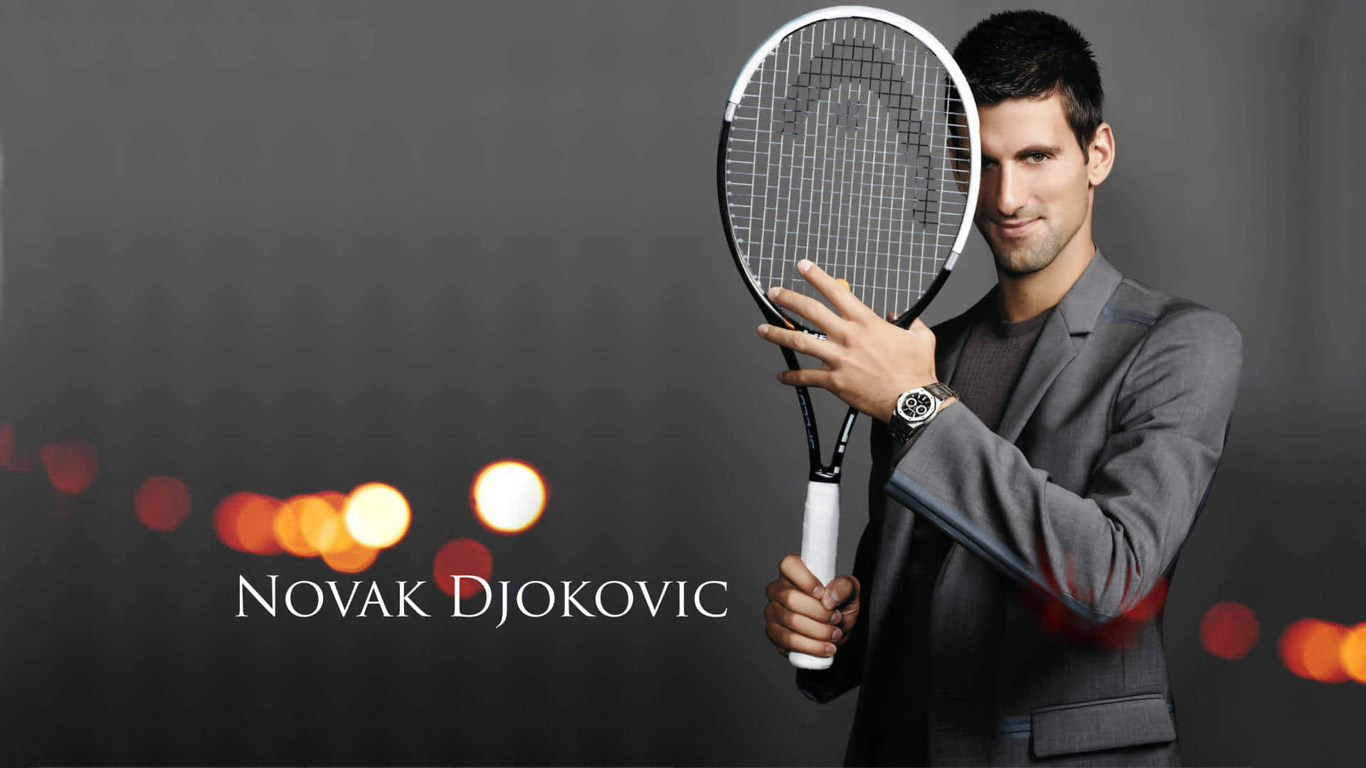 HD Tennis 桌布 壁紙 — 諾瓦克·喬科維奇身穿俐落灰色西裝，在黑暗背景下手持網球拍，背景有溫暖的散景燈光。