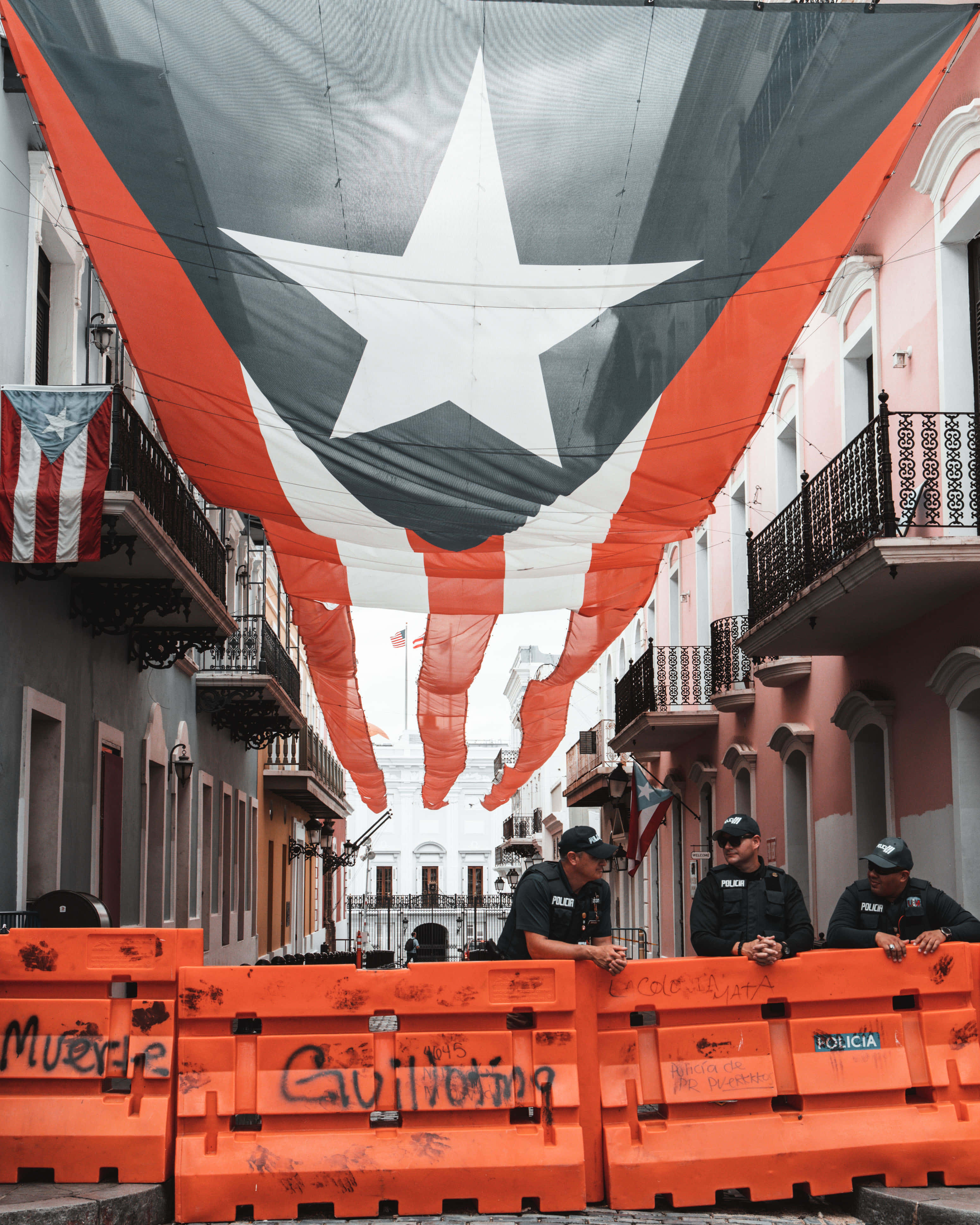 Puerto Rican 桌布 壁紙 — 警察站在橙色路障後面，上方懸掛著一面巨大的波多黎各國旗，旗幟垂落在殖民時期建築之間。
