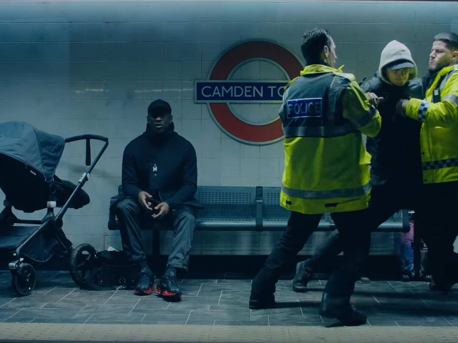 Skepta 桌布 壁紙 — 警察護送一名戴頭套的人穿過光線昏暗的卡姆登鎮地鐵站，附近一名男子獨自坐著，旁邊有一輛嬰兒車。