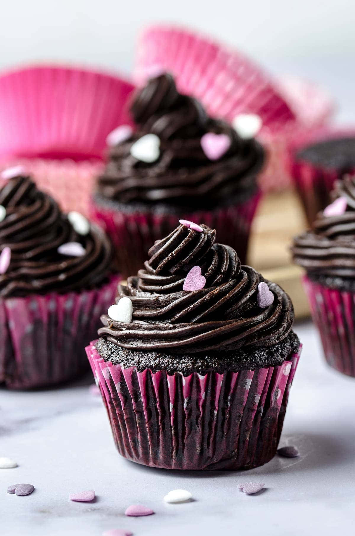 Cupcake 桌布 壁紙 — 豐富的巧克力杯子蛋糕放在粉紅色紙托上，頂部有深色糖霜漩渦和心形糖粒，置於大理石表面。