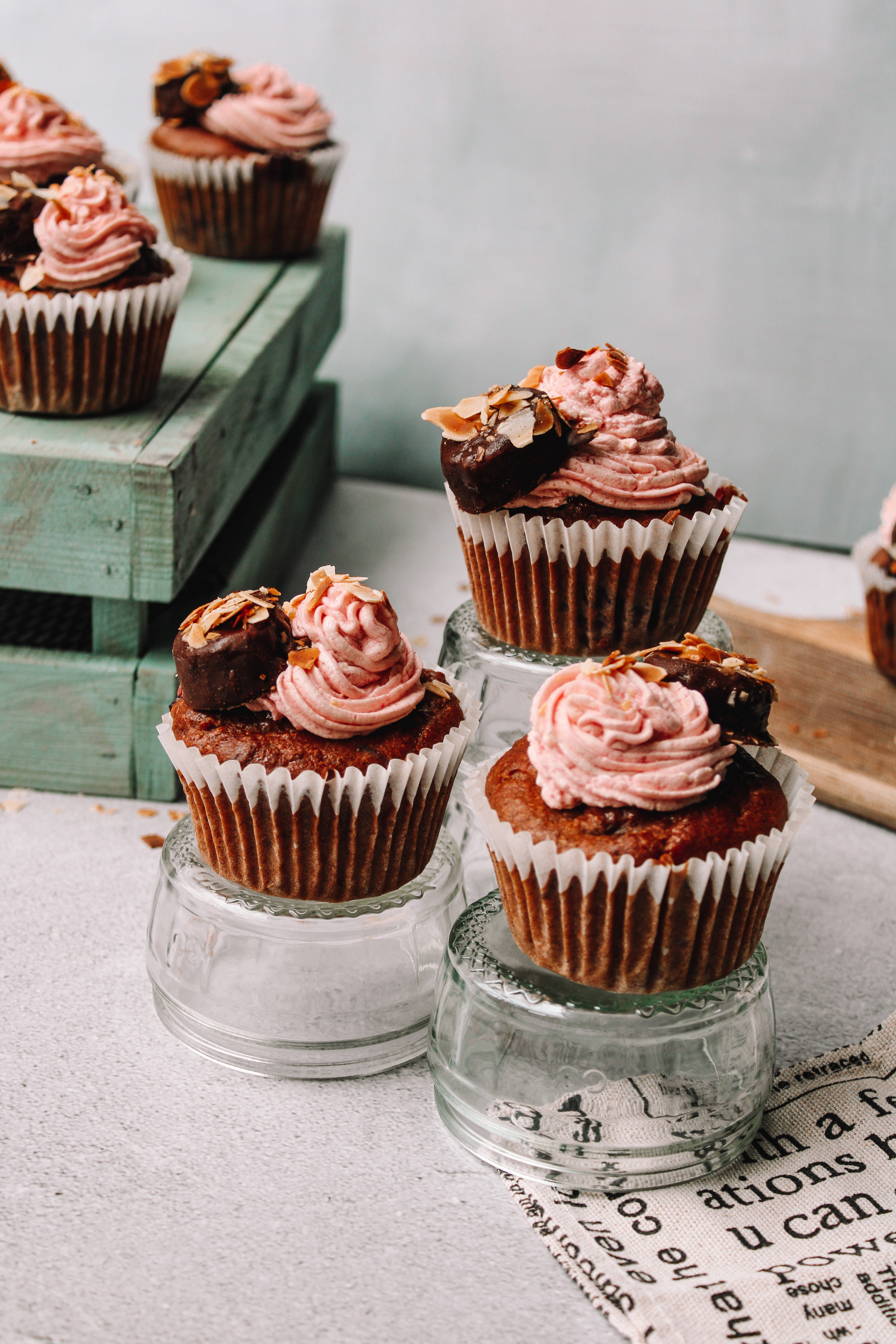Cupcake 桌布 壁紙 — 豐富的巧克力杯子蛋糕配上粉紅色糖霜和杏仁裝飾，展示在玻璃架上，背景是柔和的綠色木箱。