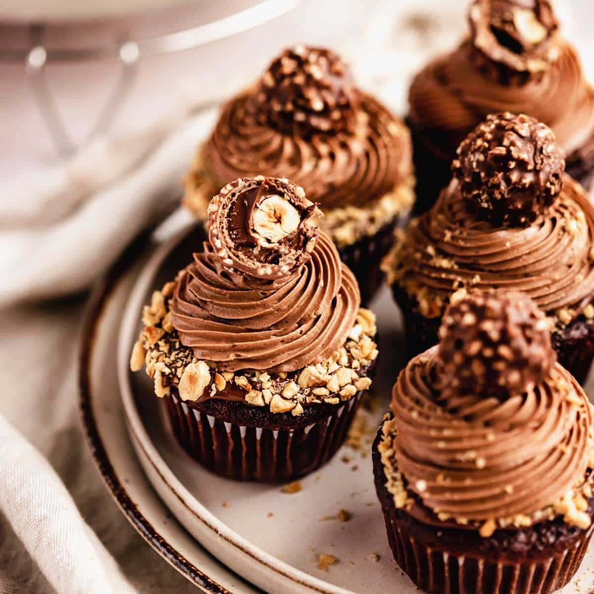 Cupcake 桌布 壁紙 — 豐富的巧克力紙杯蛋糕，上面有糖霜漩渦和碎榛子，放在陶瓷盤上。