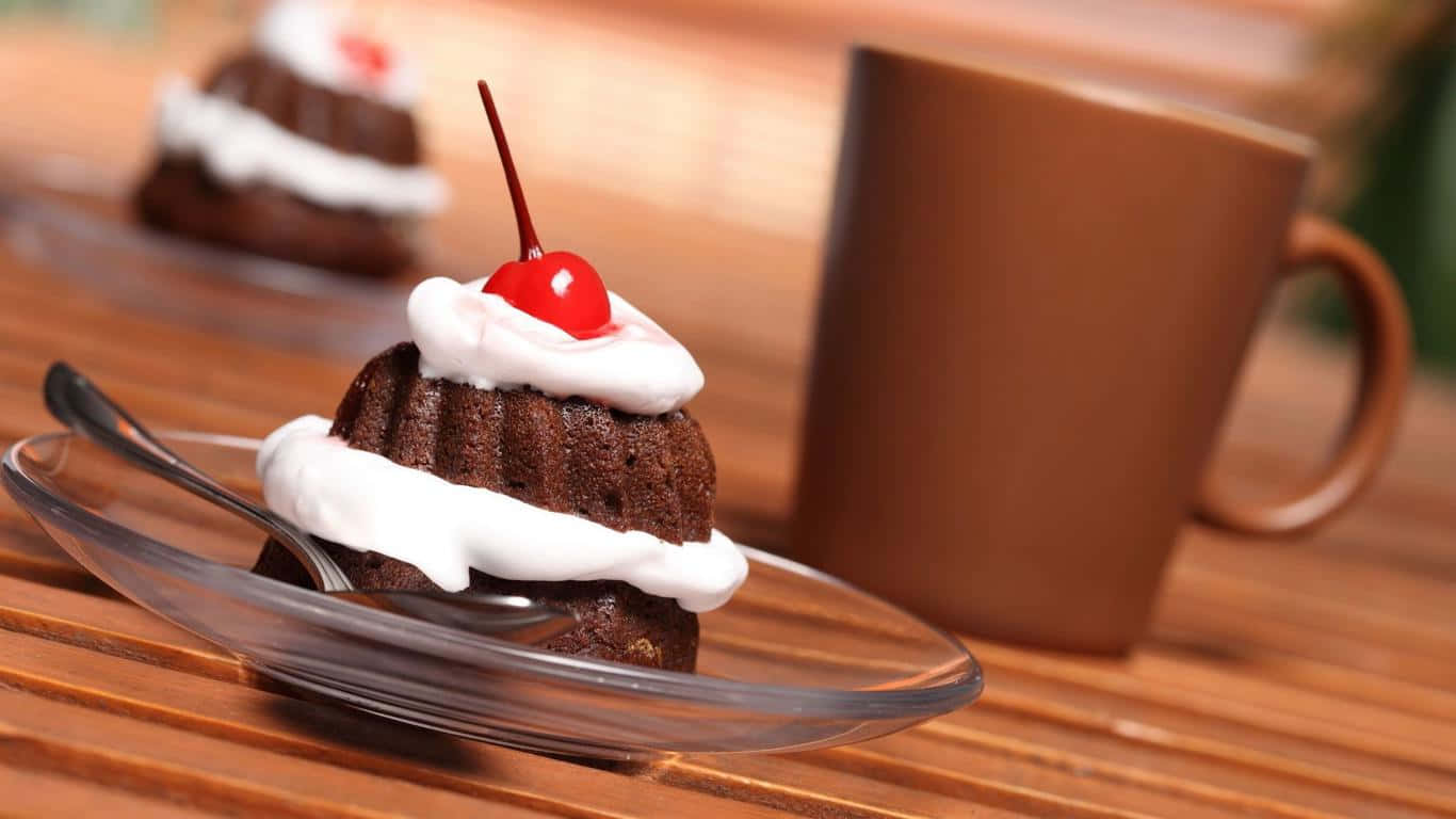 1366x768 Desserts 桌布 壁紙 — 豐富的巧克力迷你蛋糕，上面有鮮奶油和鮮紅櫻桃，放在玻璃盤上，旁邊有一個棕色馬克杯在木桌上。