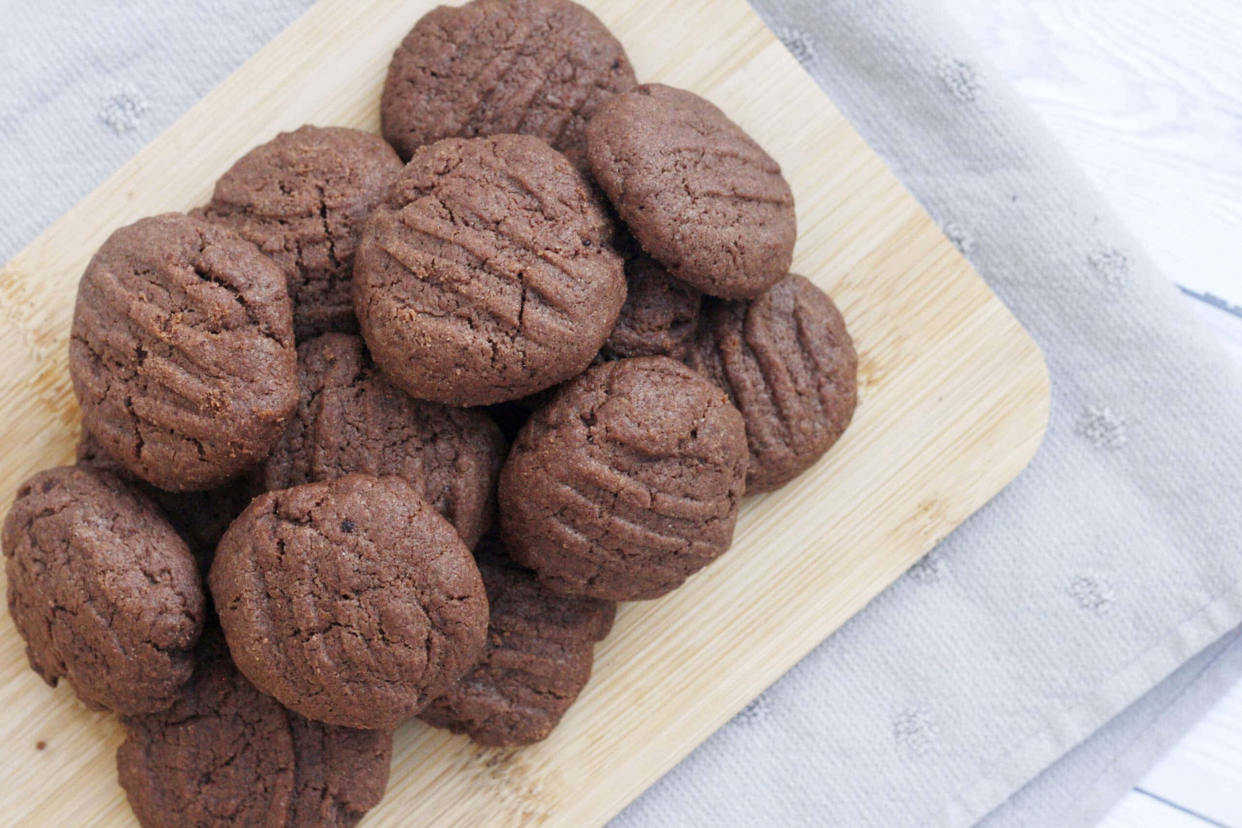 Biscuit 桌布 壁紙 — 豐富的巧克力餅乾，頂部有裂紋，堆疊在淺色木板上，置於有紋理的灰色布料上。