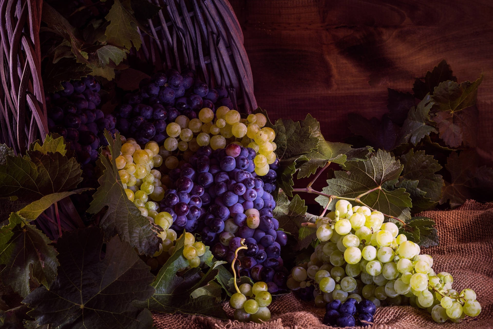 Grape 桌布 壁紙 — 豐富的紫色和綠色葡萄串從編織籃子溢出到麻布上，帶有深色陰影和溫暖的燈光。