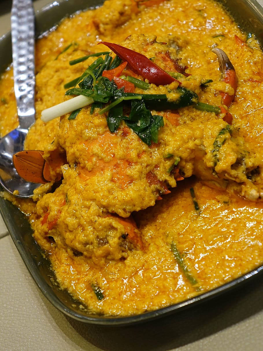 yummy 桌布 壁紙 — 豐富的金黃色蟹肉咖哩，配上紅辣椒和綠色香草，在深色碗中閃閃發光，旁邊放著一隻湯匙。