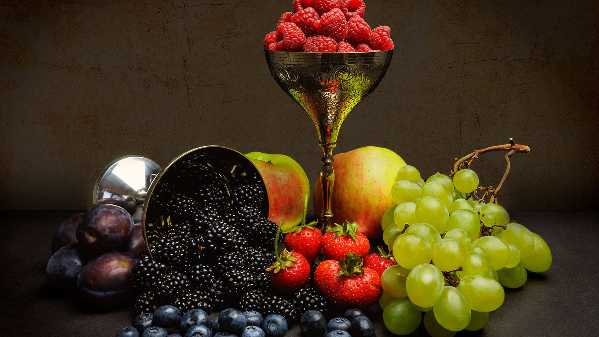 Fruit 桌布 壁紙 — 豐富的靜物畫，充滿活力的漿果、葡萄、蘋果和李子圍繞著一個華麗的高腳杯，背景有紋理。