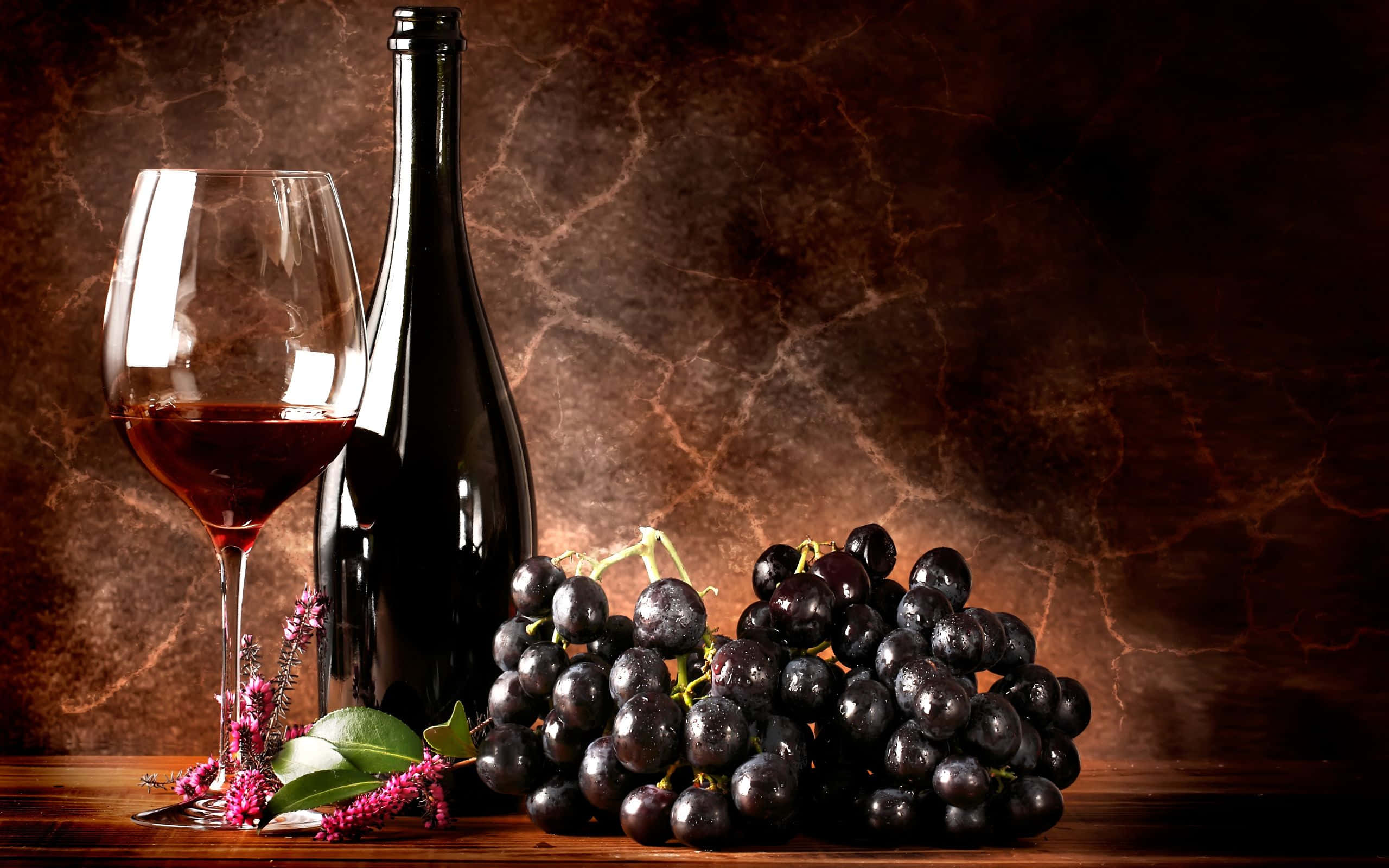 Red Wine Glass 桌布 壁紙 — 豐富的靜物畫，描繪一個玻璃瓶和一杯紅酒，旁邊是一串深色葡萄，放在木桌上，背景是帶紋理的棕色背景。