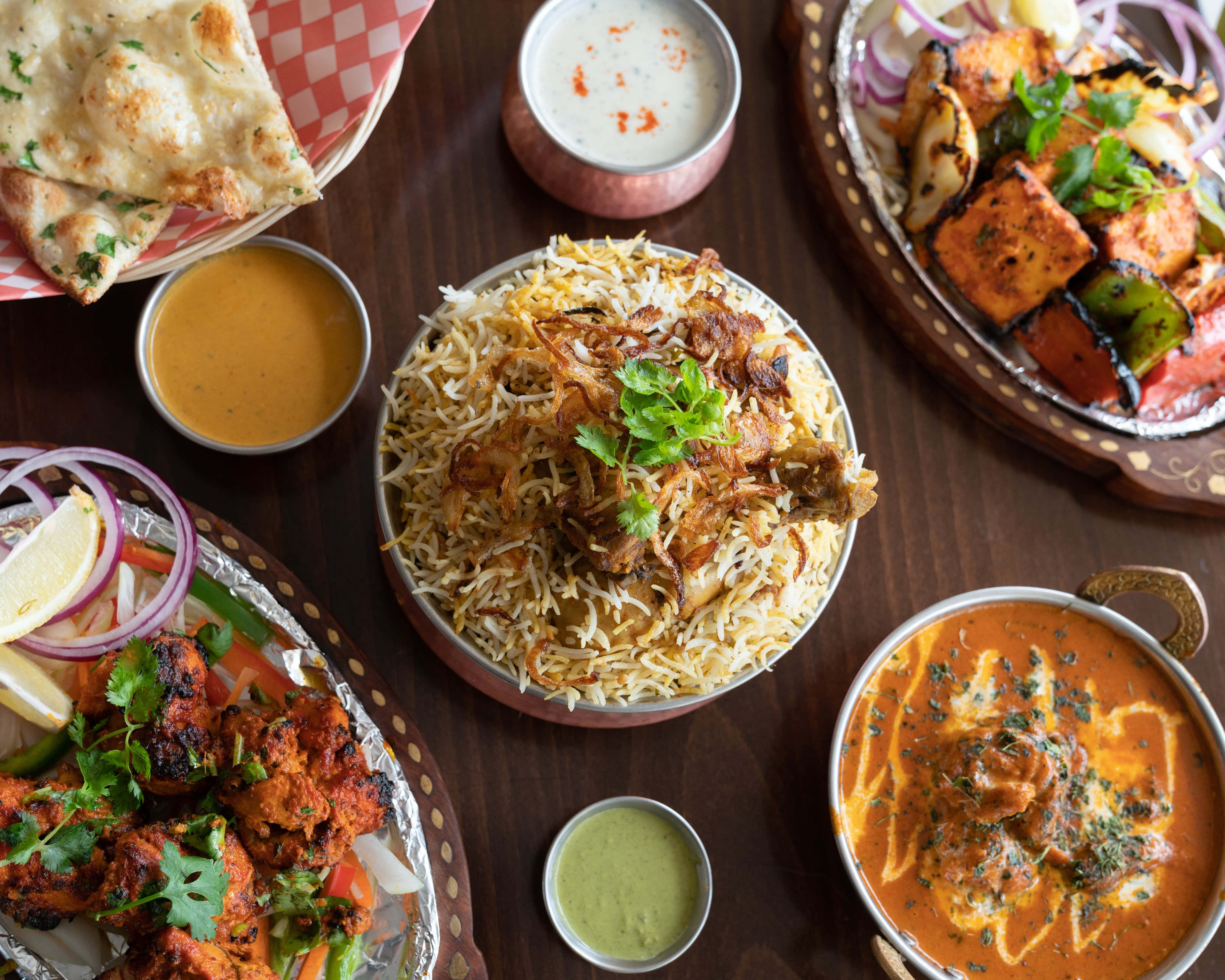 Indian 桌布 壁紙 — 豐盛的印度菜餚，包括印度香飯雞肉、烤串、咖哩、烤餅和色彩繽紛的酸辣醬，擺放在深色木桌上。