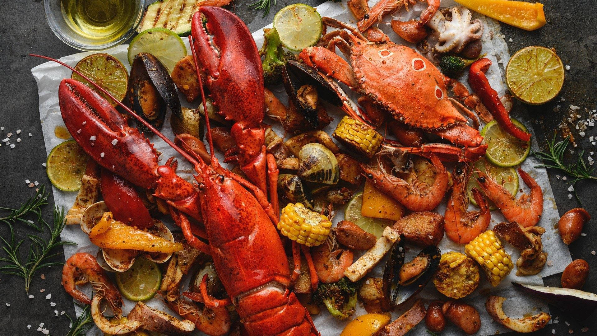 Lobster 桌布 壁紙 — 豐盛的海鮮煮物，包括鮮紅龍蝦、螃蟹、蝦和玉米，置於深色紋理表面上，以萊姆和香草點綴。