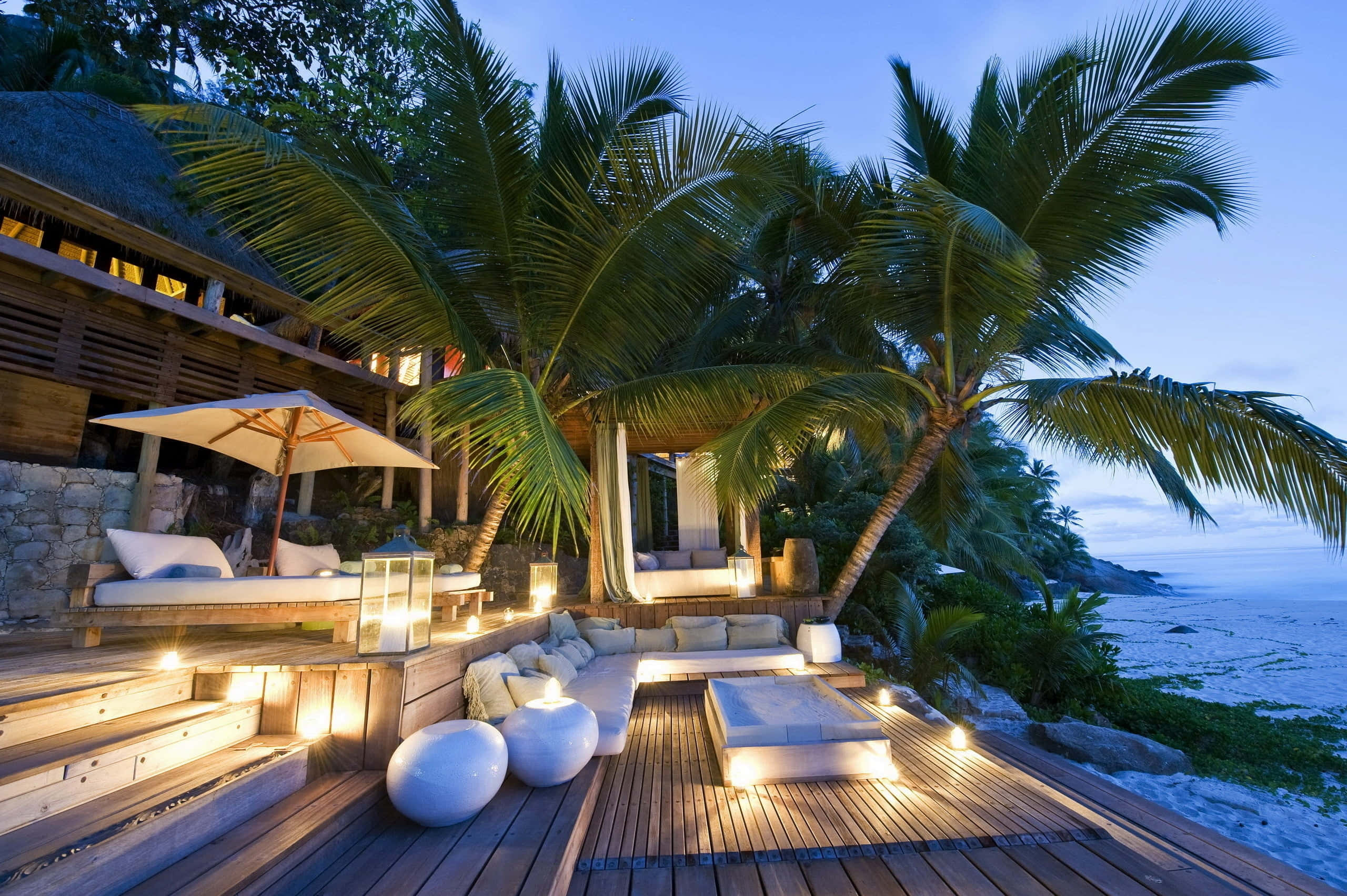 Beach Daybed 桌布 壁紙 — 豪華木甲板，在棕櫚樹下，黃昏時分的海邊度假村，配有發光燈籠和白色躺椅家具。