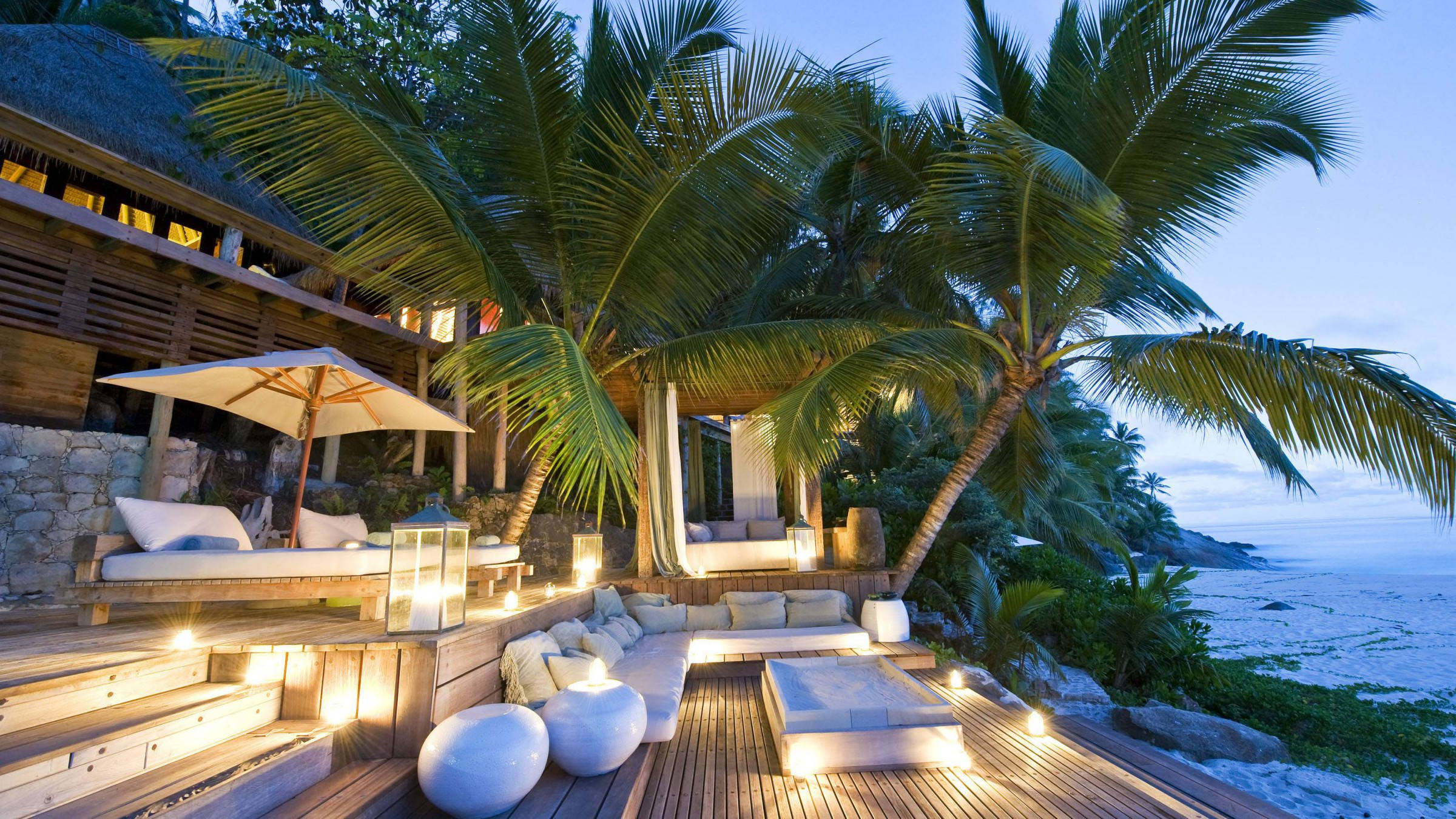 Seychelles 桌布 壁紙 — 豪華木甲板，在沙灘旁的暮色中，棕櫚樹下閃爍的燈籠和白色躺椅家具。