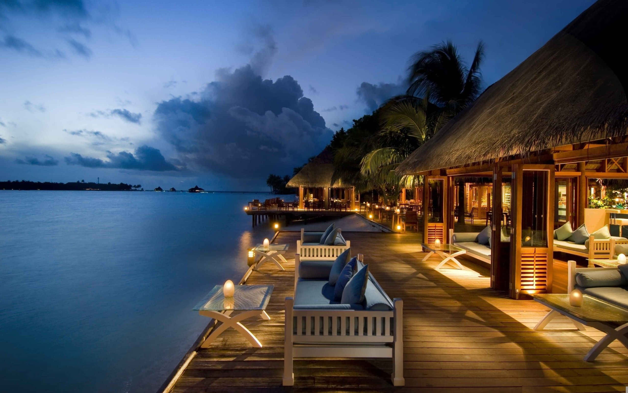 Beach Resort 桌布 壁紙 — 豪華水上平房在黃昏時分，在戲劇性的雲層下平靜的海水旁溫暖地發光。