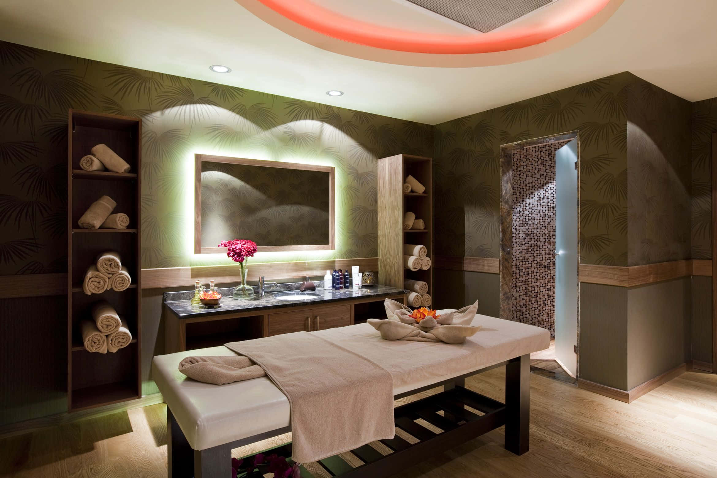 Massage Room 桌布 壁紙 — 豪華水療室，配有木製按摩床、發光天花板環和棕櫚圖案牆壁，沐浴在溫暖的環境光中。