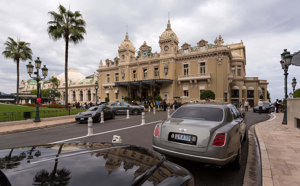 Luxury 桌布 壁紙 — 豪華汽車排列在宏偉的蒙地卡羅賭場前的街道上，天空多雲，棕櫚樹點綴其間。