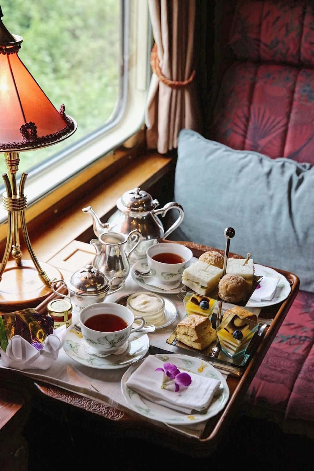 Teatime 桌布 壁紙 — 豪華火車車廂茶具套裝，配有銀器、糕點和一盞發光的燈，旁邊是窗外有綠植的窗戶。