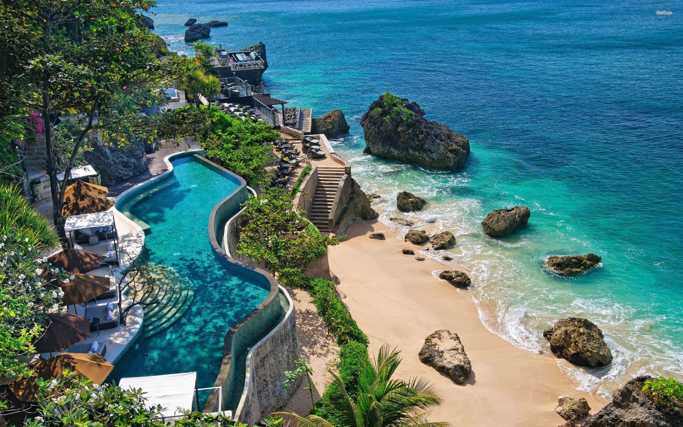 Bali Beach 桌布 壁紙 — 豪華無邊際泳池沿著懸崖傾瀉而下，旁邊是沙灘、碧綠海水和散落的岩石。