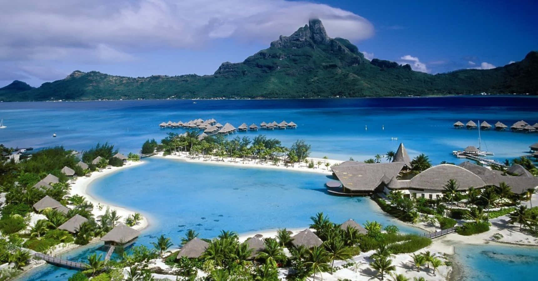 Islands 桌布 壁紙 — 豪華熱帶度假村的鳥瞰圖，水上平房坐落在碧綠的潟湖中，周圍環繞著鬱鬱蔥蔥的綠色山脈，在明亮的藍天下。