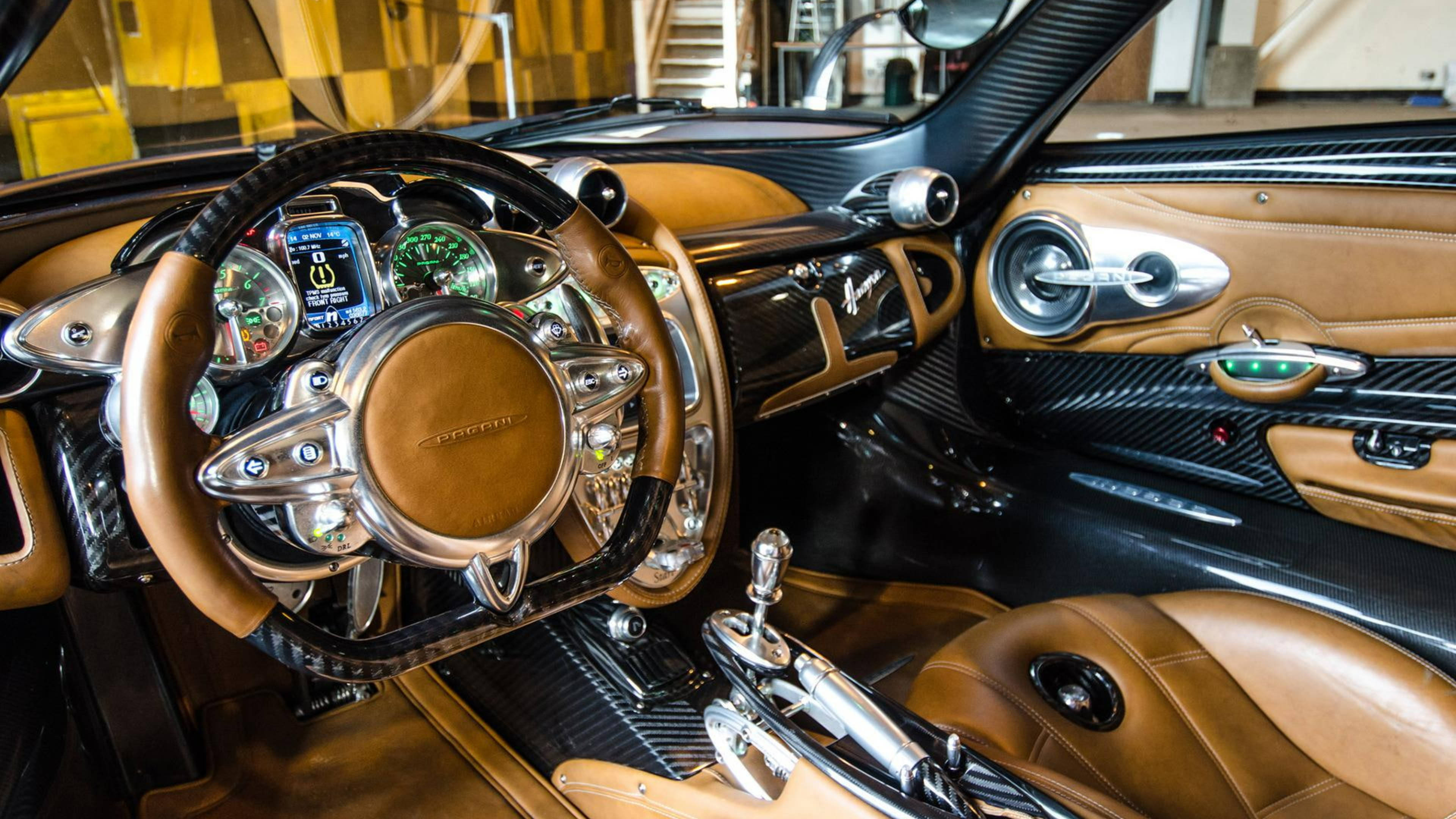 Pagani 桌布 壁紙 — 豪華車內裝，配備棕褐色皮革座椅和碳纖維飾邊，在明亮的車庫燈光下。