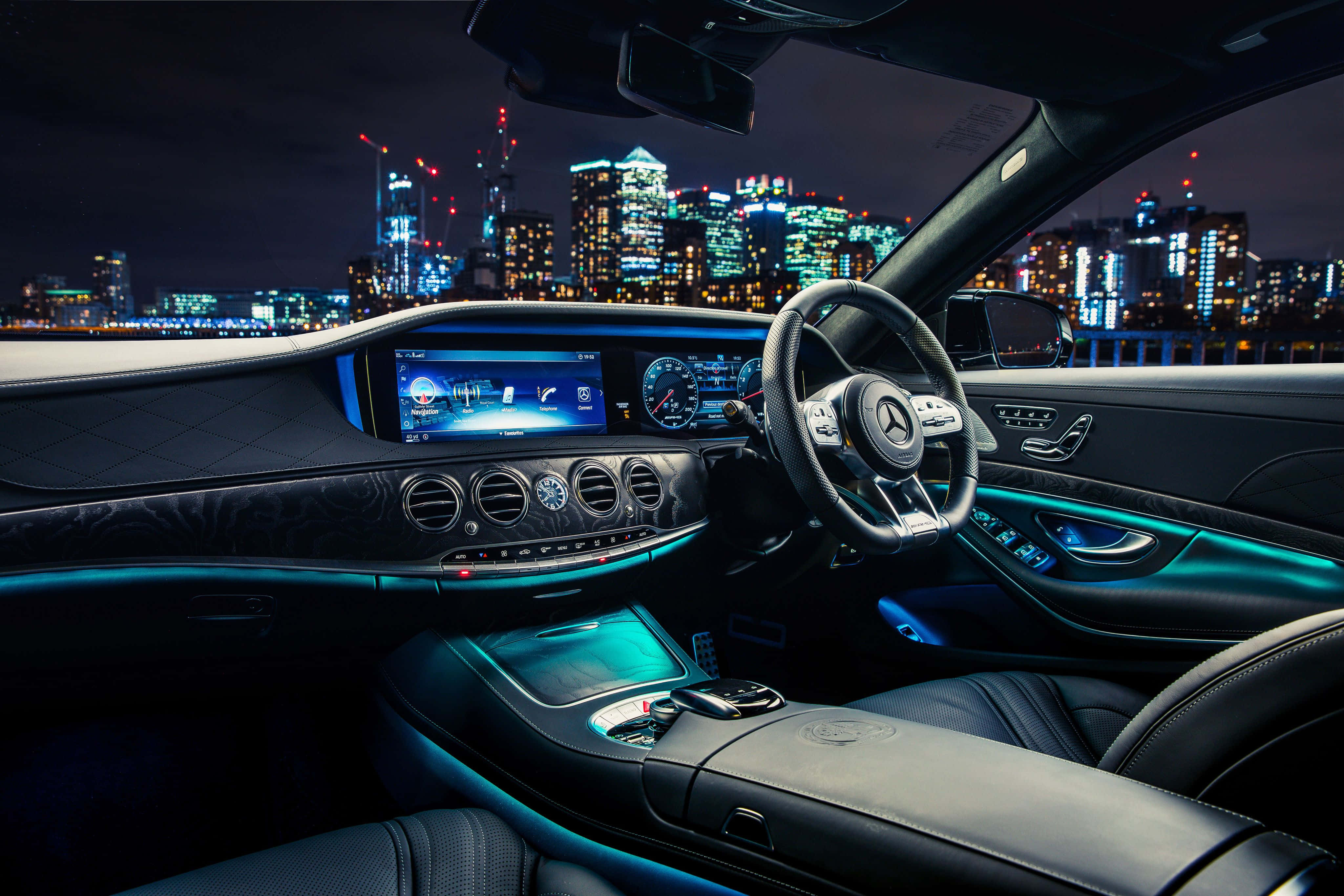 Mercedes Car 4K 桌布 壁紙 — 豪華車內裝在夜晚，帶有發光的藍色氛圍燈和透過擋風玻璃可見的充滿活力的城市天際線。
