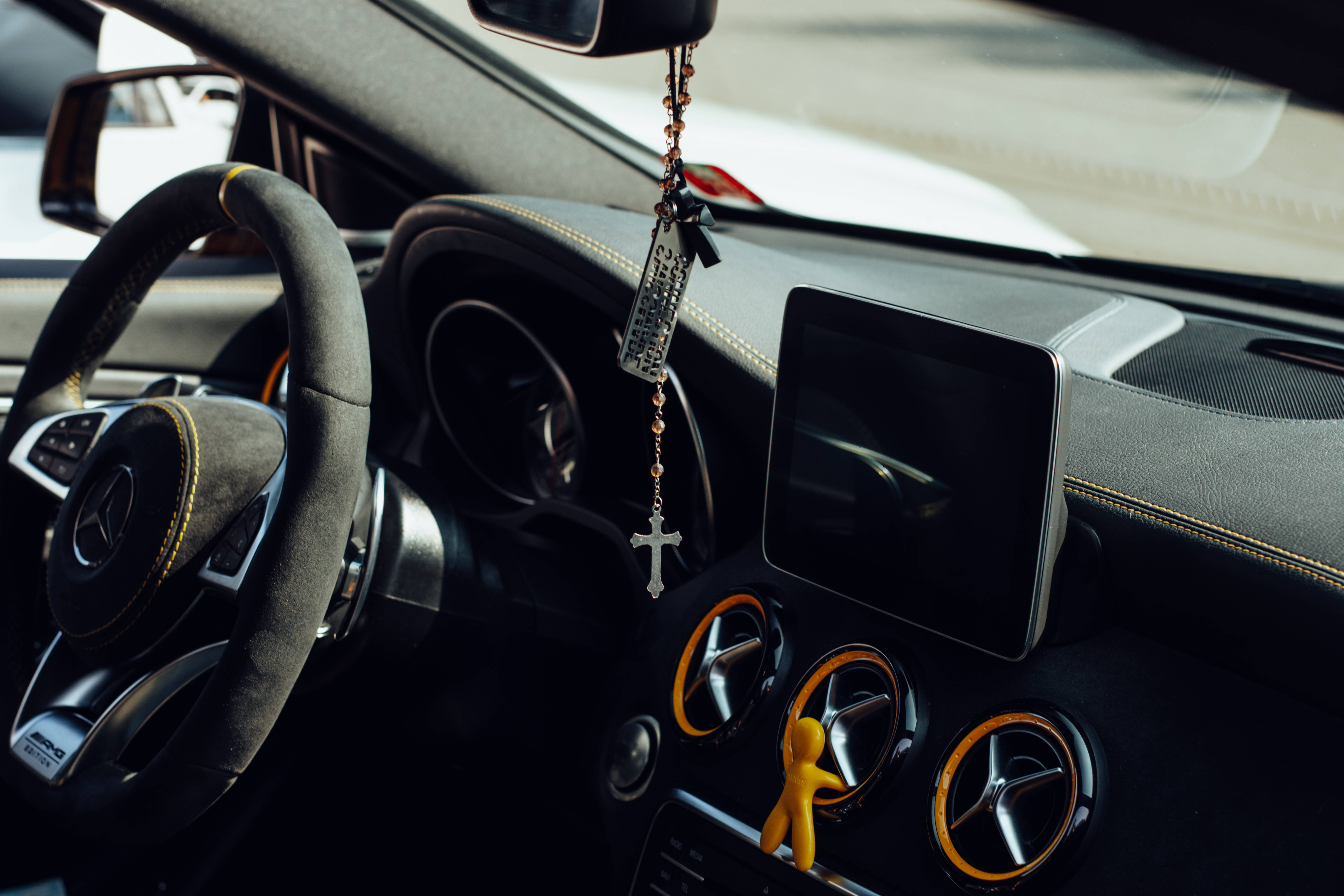 Mercedes Amg 桌布 壁紙 — 豪華車內飾特寫，顯示方向盤、掛在後視鏡上的玫瑰經念珠和儀表板上的黃色公仔。