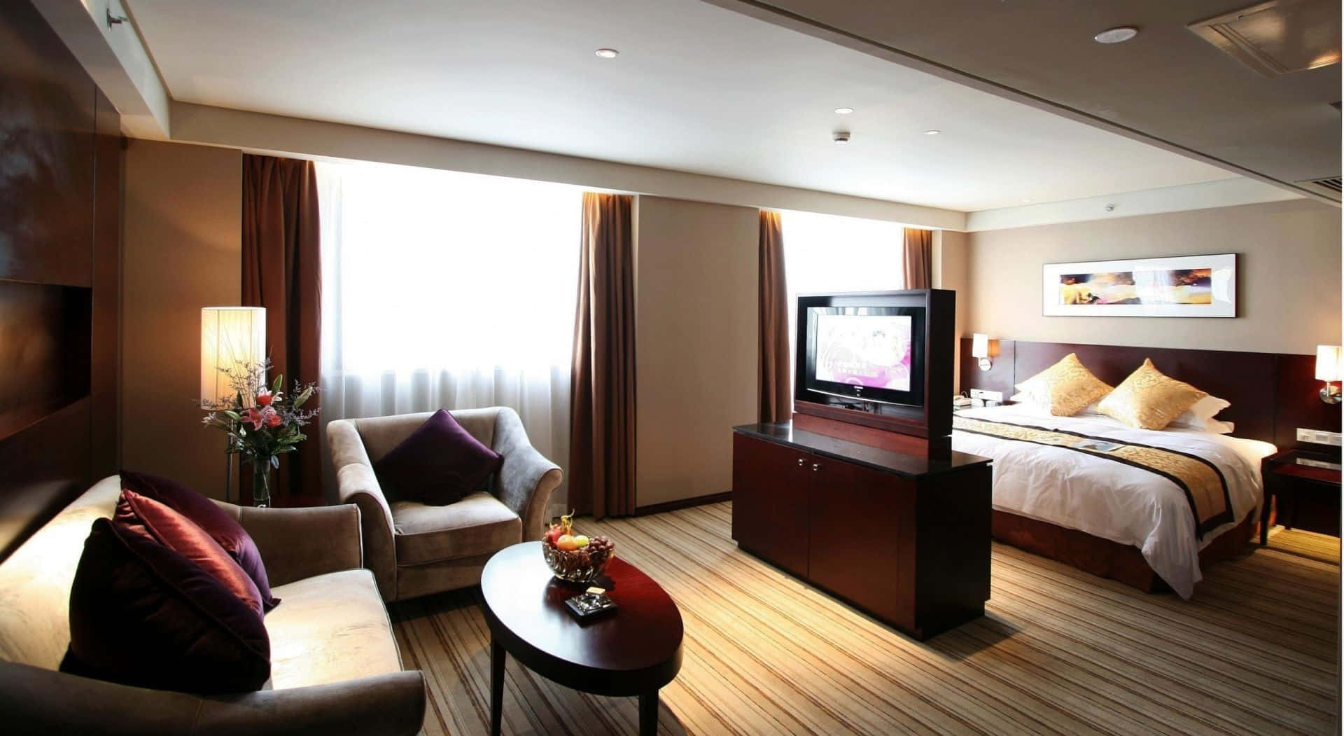 Hotel Room 桌布 壁紙 — 豪華酒店客房，鋪有木地板，配備一張大床和一個舒適的休息區，沐浴在柔和自然光中。