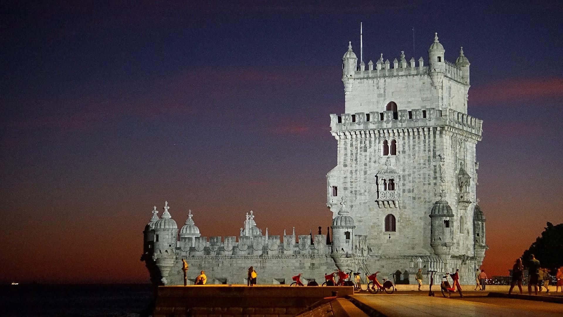 Portugal 桌布 壁紙 — 貝倫塔在暮色中亮起，暖橙色與深藍色交織，人們沿著海濱漫步。