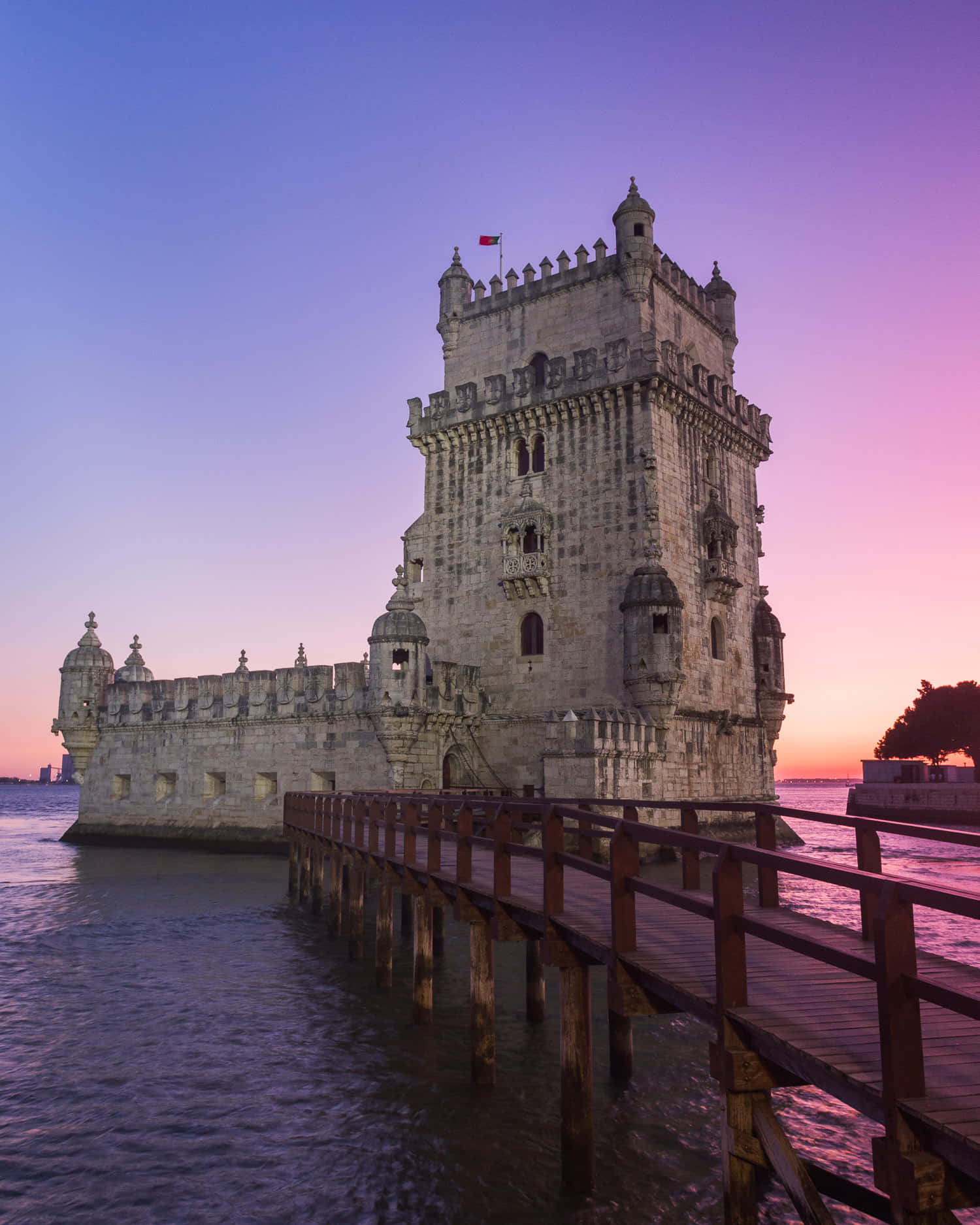 Portugal 桌布 壁紙 — 貝倫塔在黃昏時分巍然聳立，粉紅色和紫色的天空倒映在平靜的水面上，木製碼頭通向其歷史悠久的石質底座。