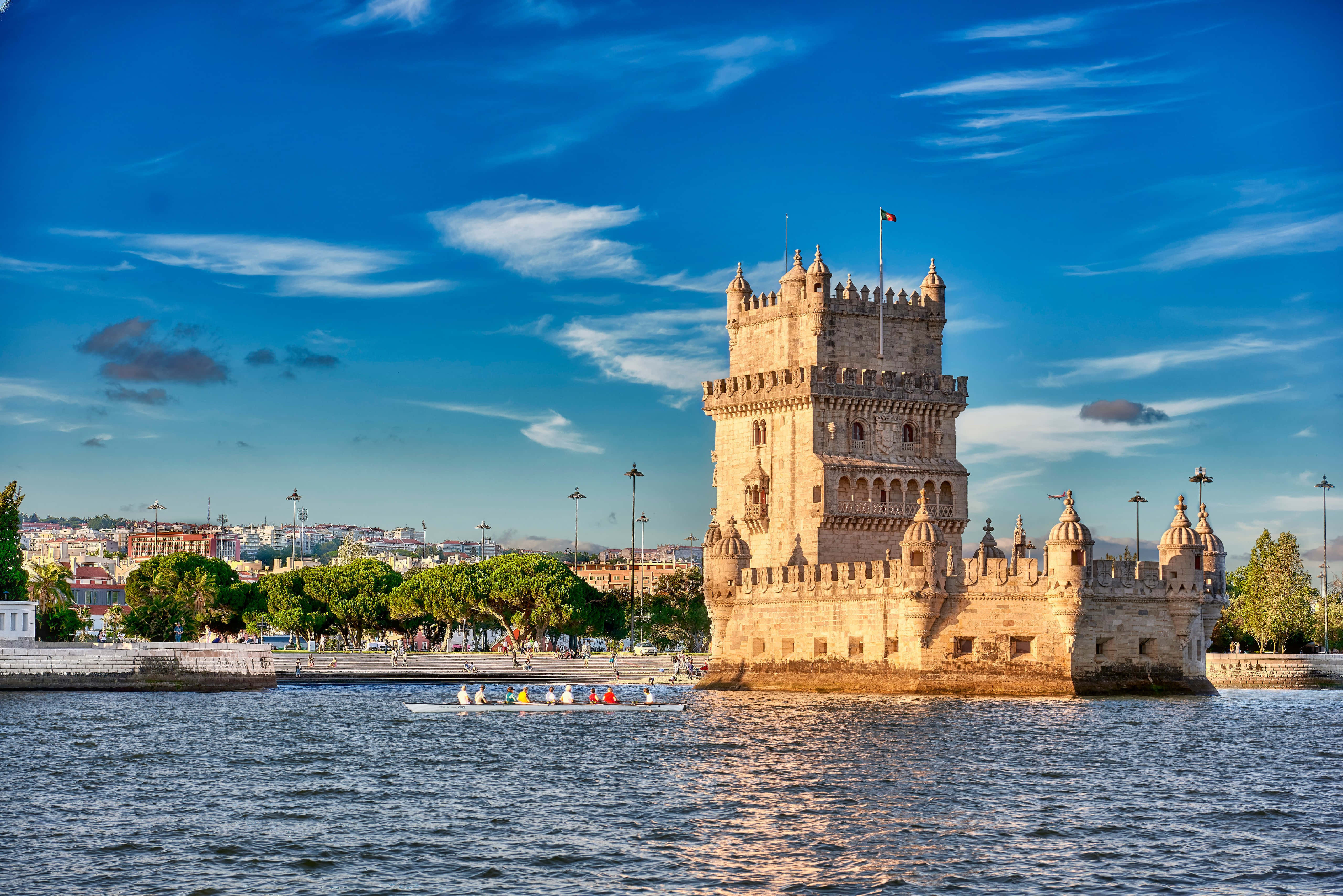 Portugal 桌布 壁紙 — 貝倫塔雄偉地矗立在水面上，天空湛藍，雲朵點綴，附近有一艘划艇滑過。