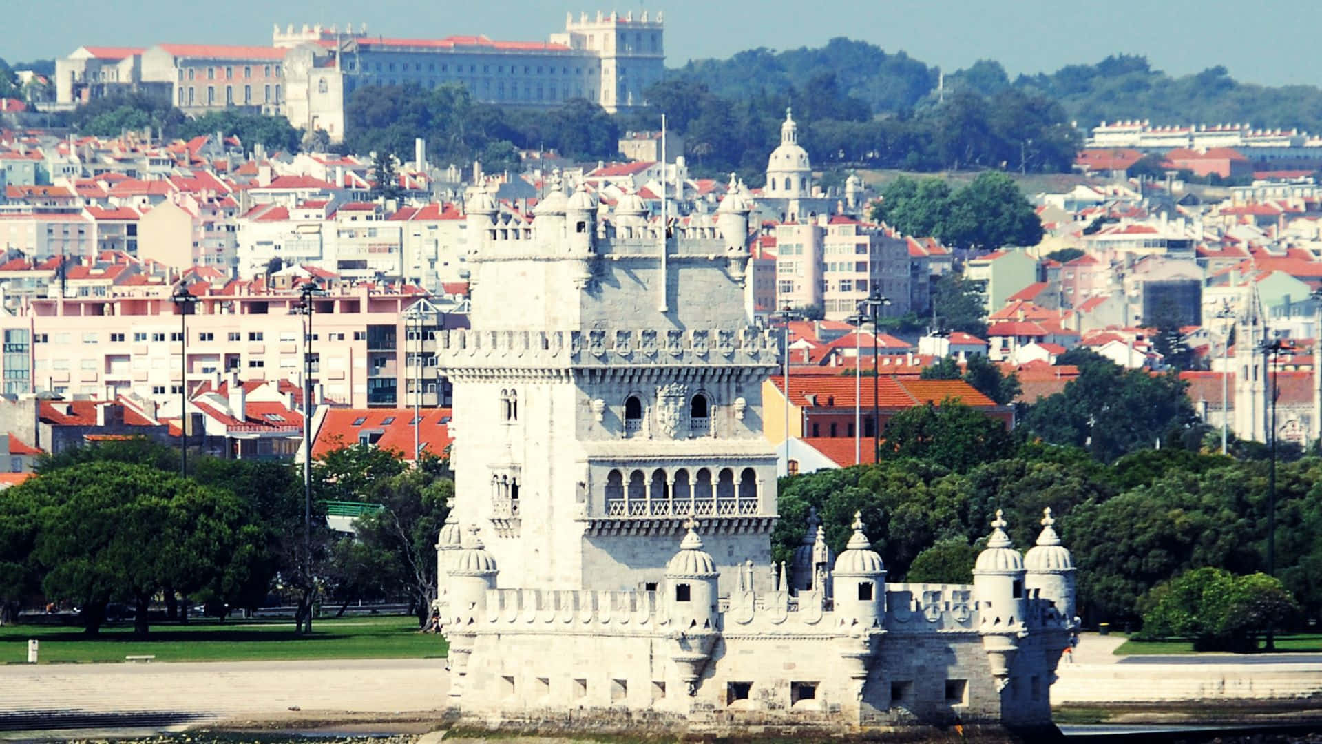 Portugal 桌布 壁紙 — 貝倫塔雄偉地矗立在河邊，里斯本的紅瓦屋頂和綠色山丘在清澈的藍天下在其後方升起。