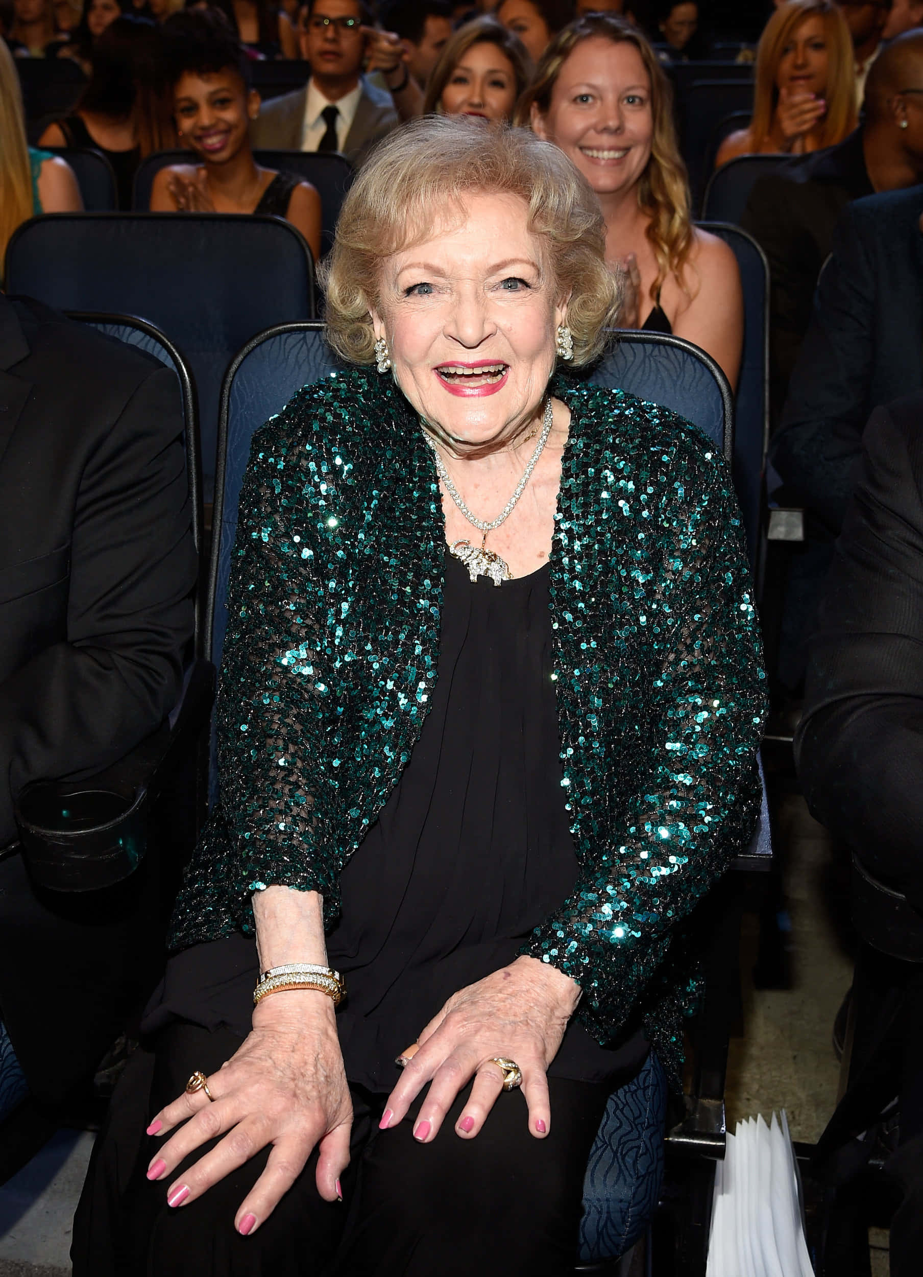 Betty White 桌布 壁紙 — 貝特·米德勒在正式活動中穿著閃閃發光的青綠色外套，光芒四射，周圍是鼓掌的與會者。
