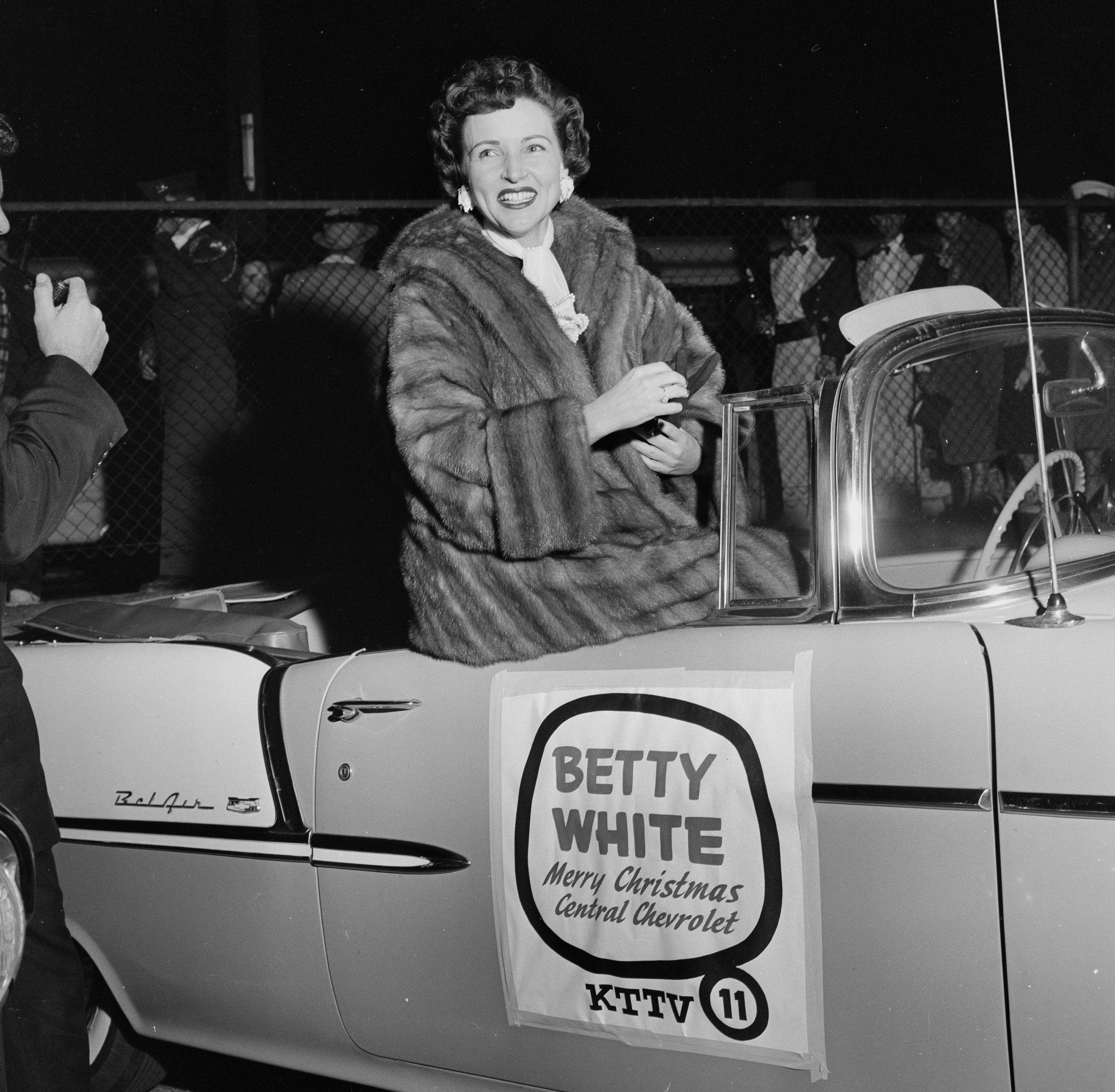 Betty White 桌布 壁紙 — 貝蒂·懷特在夜間活動中，從一輛帶有節日標誌的復古敞篷車上燦爛微笑。