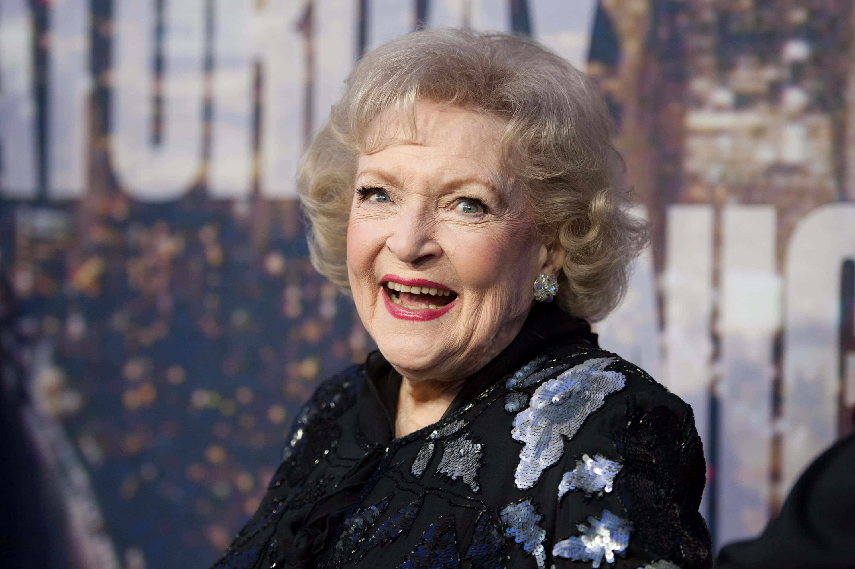 Betty White 桌布 壁紙 — 貝蒂·懷特身穿閃亮黑色亮片服裝，在模糊的電影首映背景下散發著歡樂的能量。